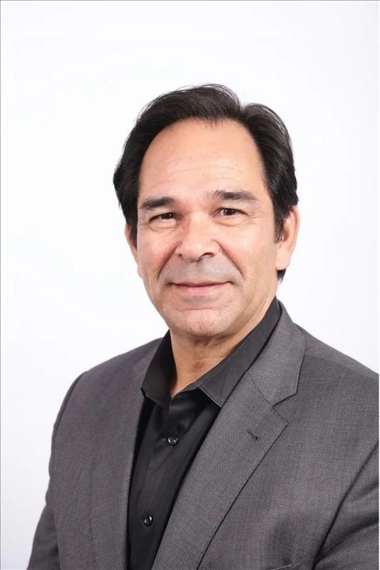 A middle-aged man with dark hair, wearing a gray suit jacket and black shirt, standing against a plain white background.
