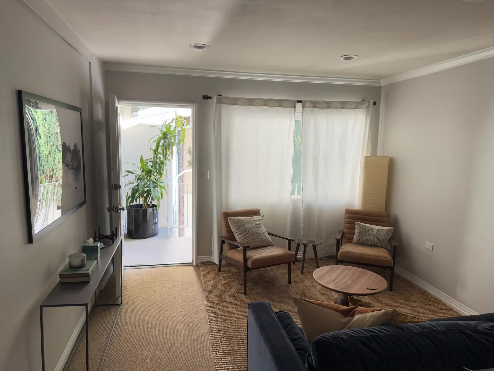 Photo of therapy office with the edge of a couch and two chairs visible. Blue velvet couch with orange pillows. Chairs are tan. Door is open with a plant visible outside the room in daylight.