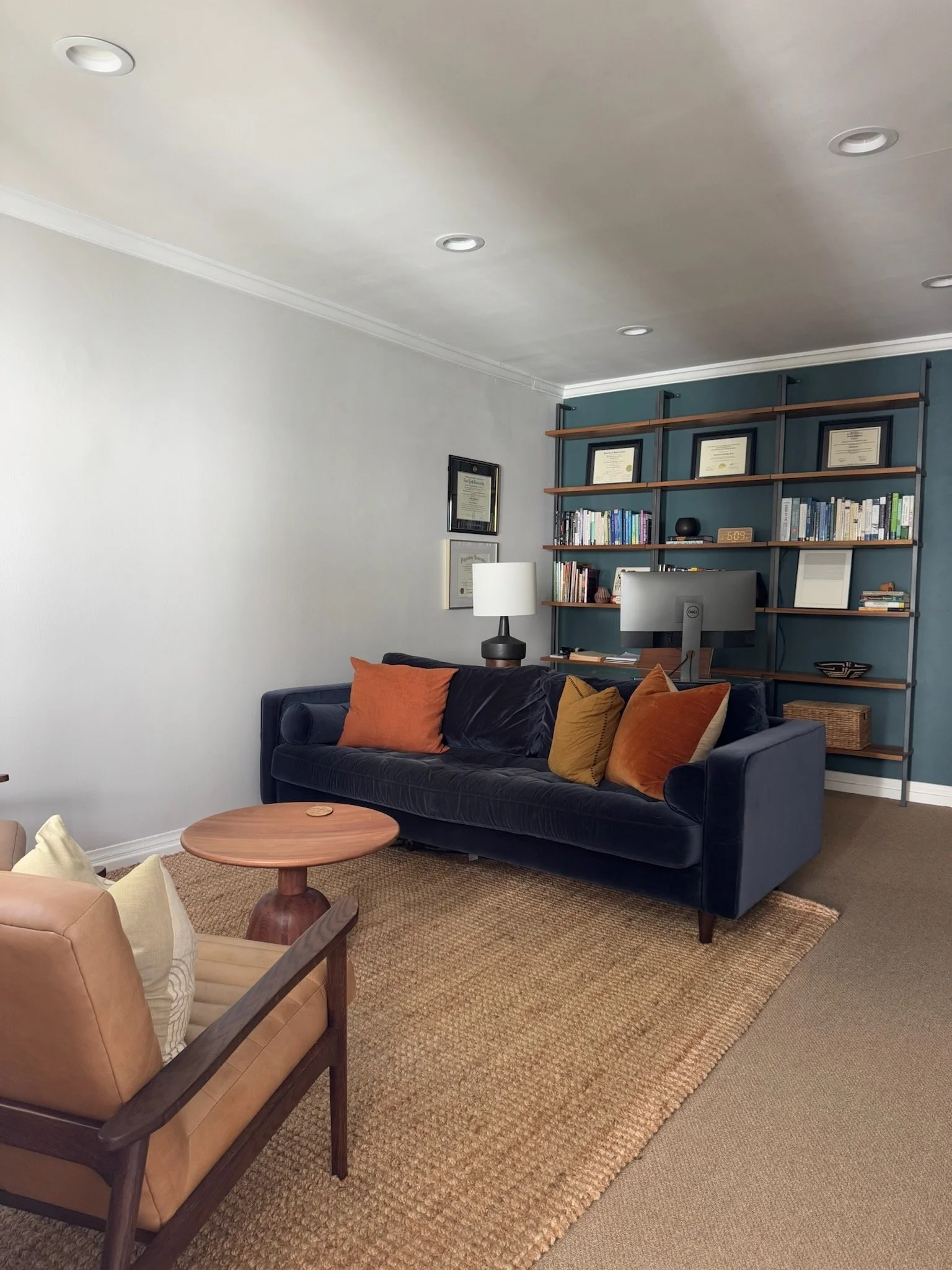 Photo of a therapy office with a comfortable couch and chairs. Blue velvet couch with orange pillows and tan chairs. Bookshelf in the background.
