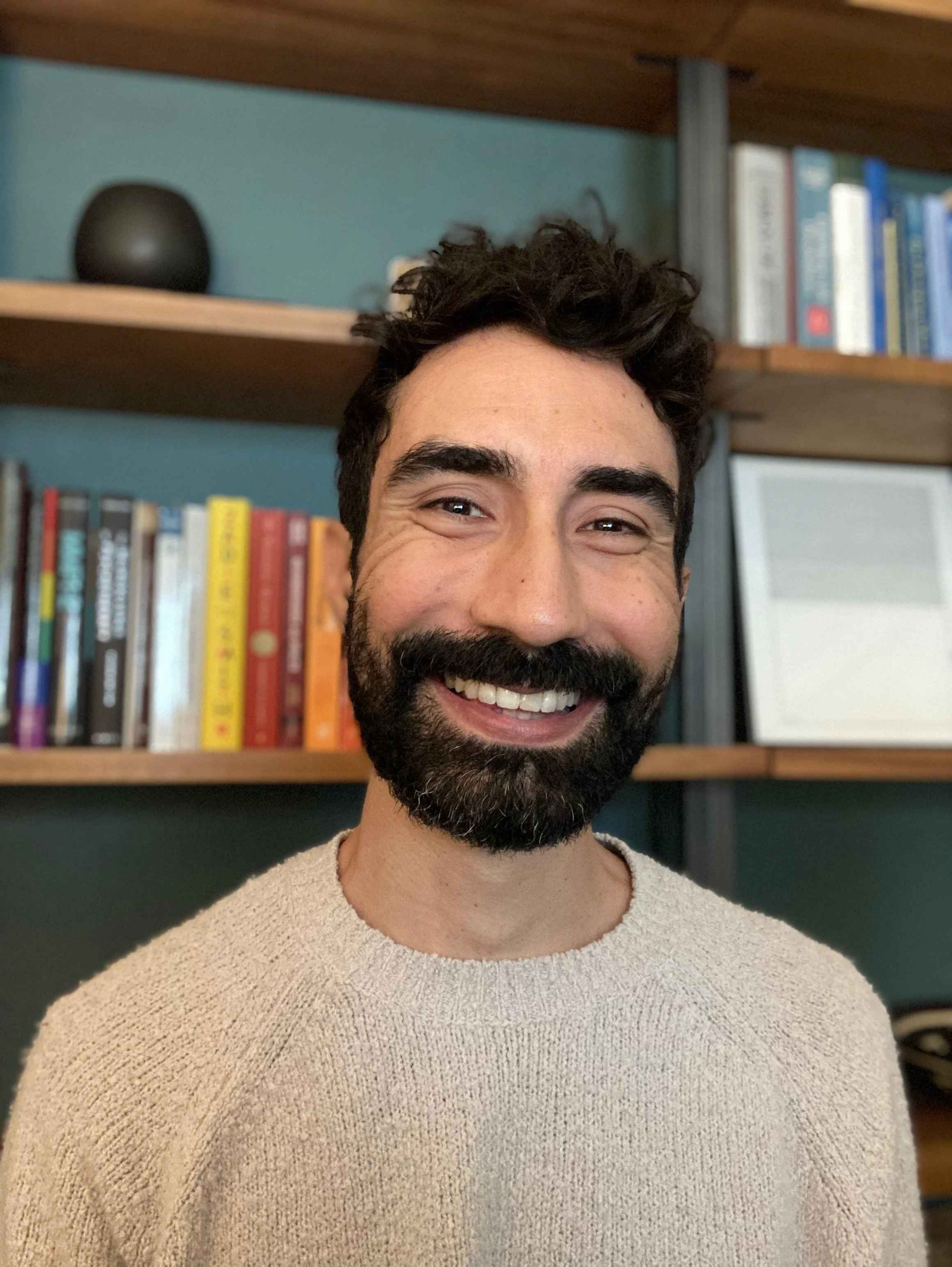 Photo of Michael Olivares PhD in his office in Los Angeles. Smiling and looking at the camera. Wearing a tan sweater in front of a bookshelf on a blue wall.