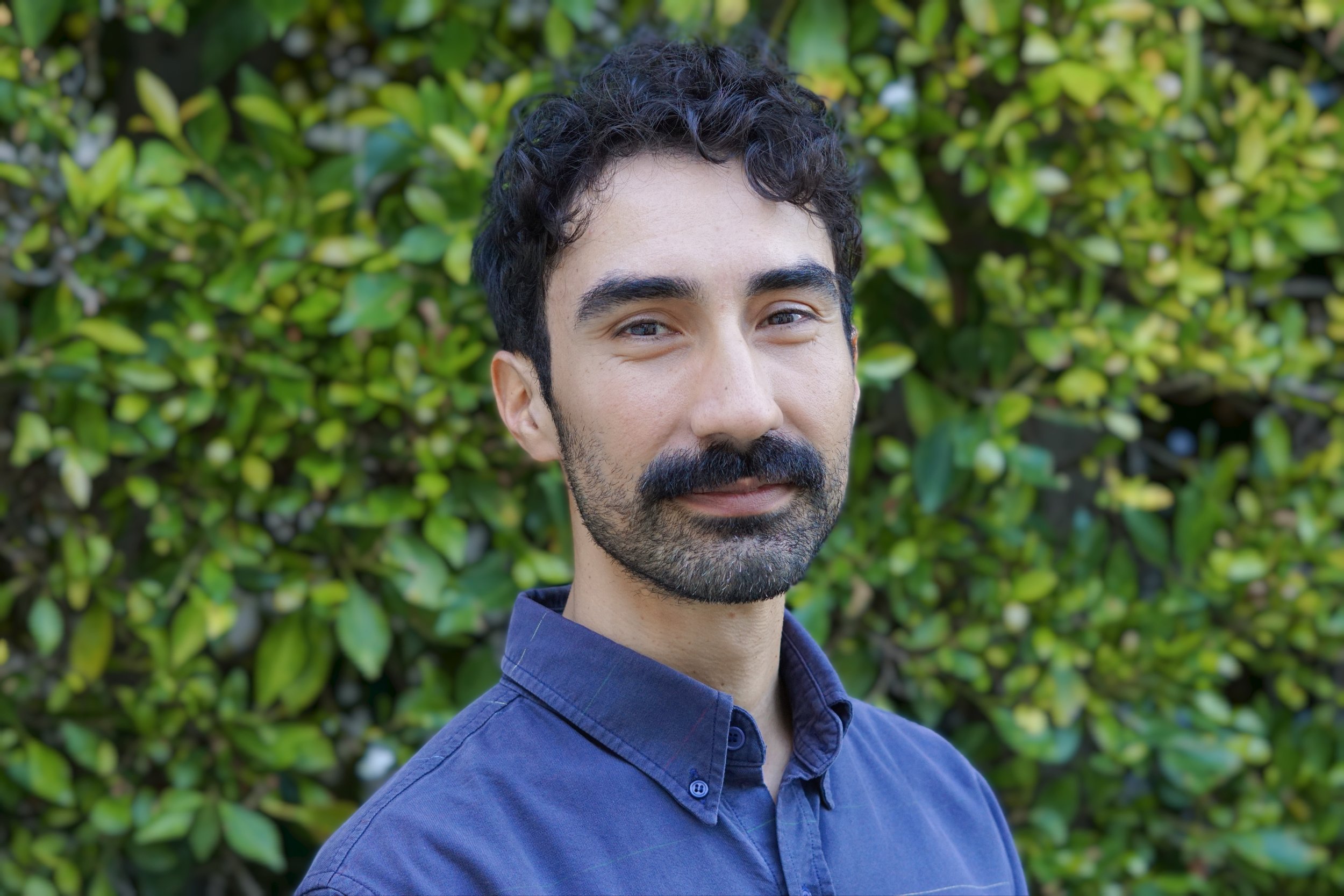 Brown multiracial male latino therapist. Smiling and welcoming. Wearing blue shirt. Green sunny leaves in the background.