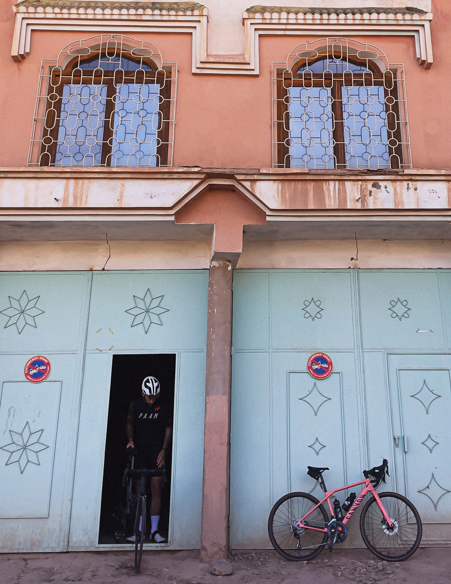 Une façade de bâtiment avec deux fenêtres ornées de grilles en fer forgé, un homme en cuissard et casque de vélo sortant d'une porte, une bicyclette rose garée contre le mur, et deux stickers en bas de la porte.