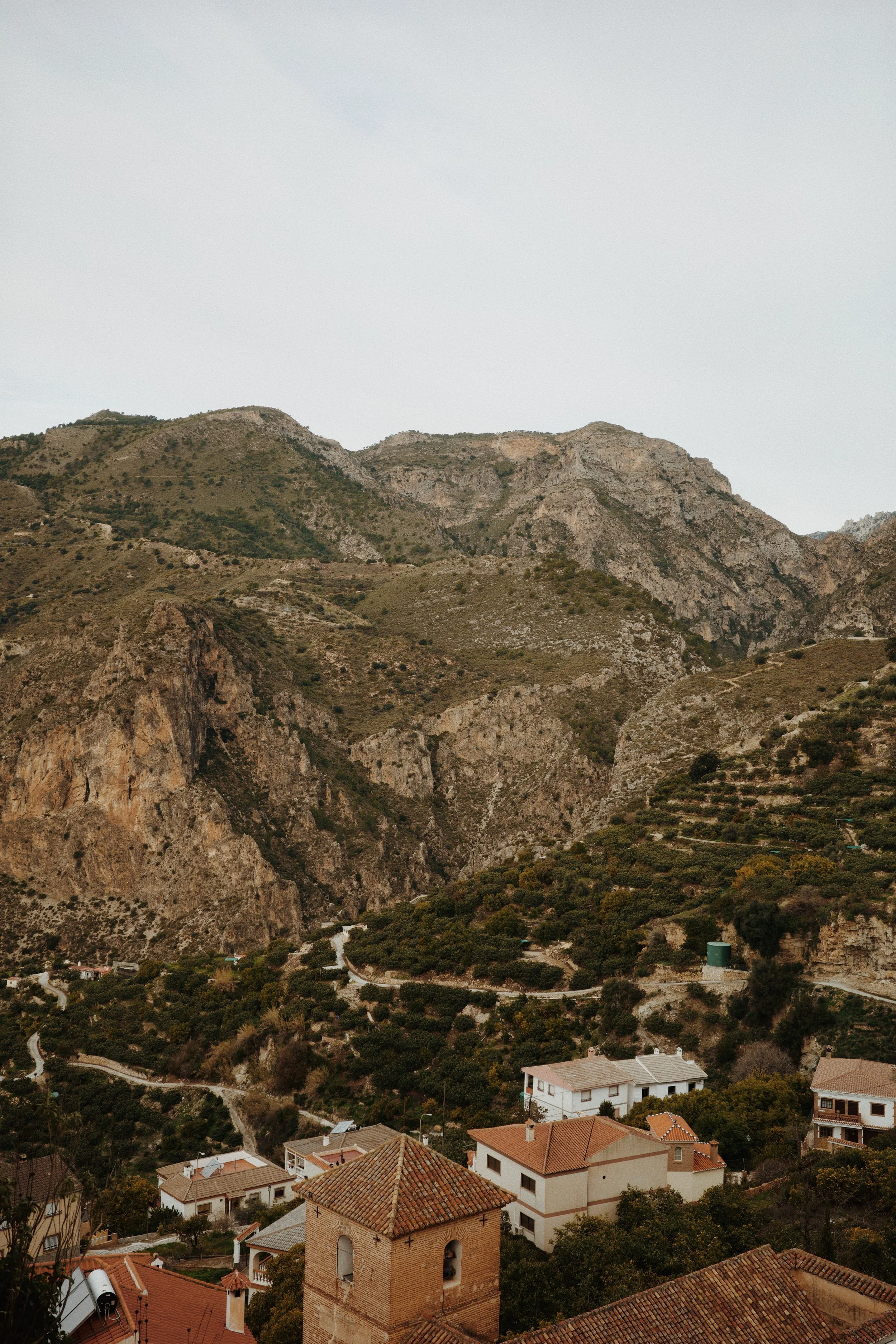 Paysage de montagnes rocheuses avec quelques maisons dans une vallée verdoyante, sous un ciel grisâtre.