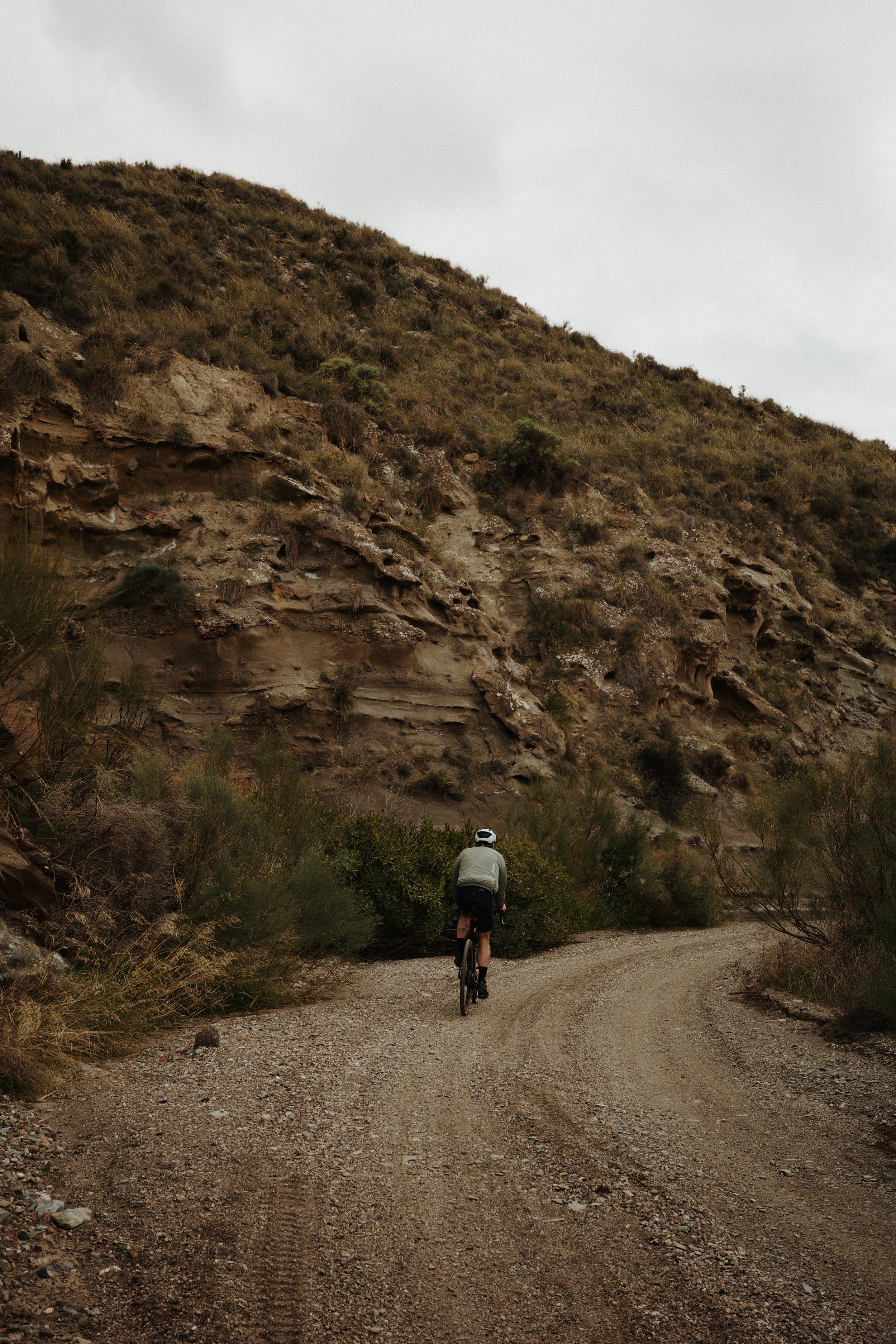 Un cycliste sur un chemin dirt dans une zone montagneuse avec des collines arides et des broussailles, ciel nuageux