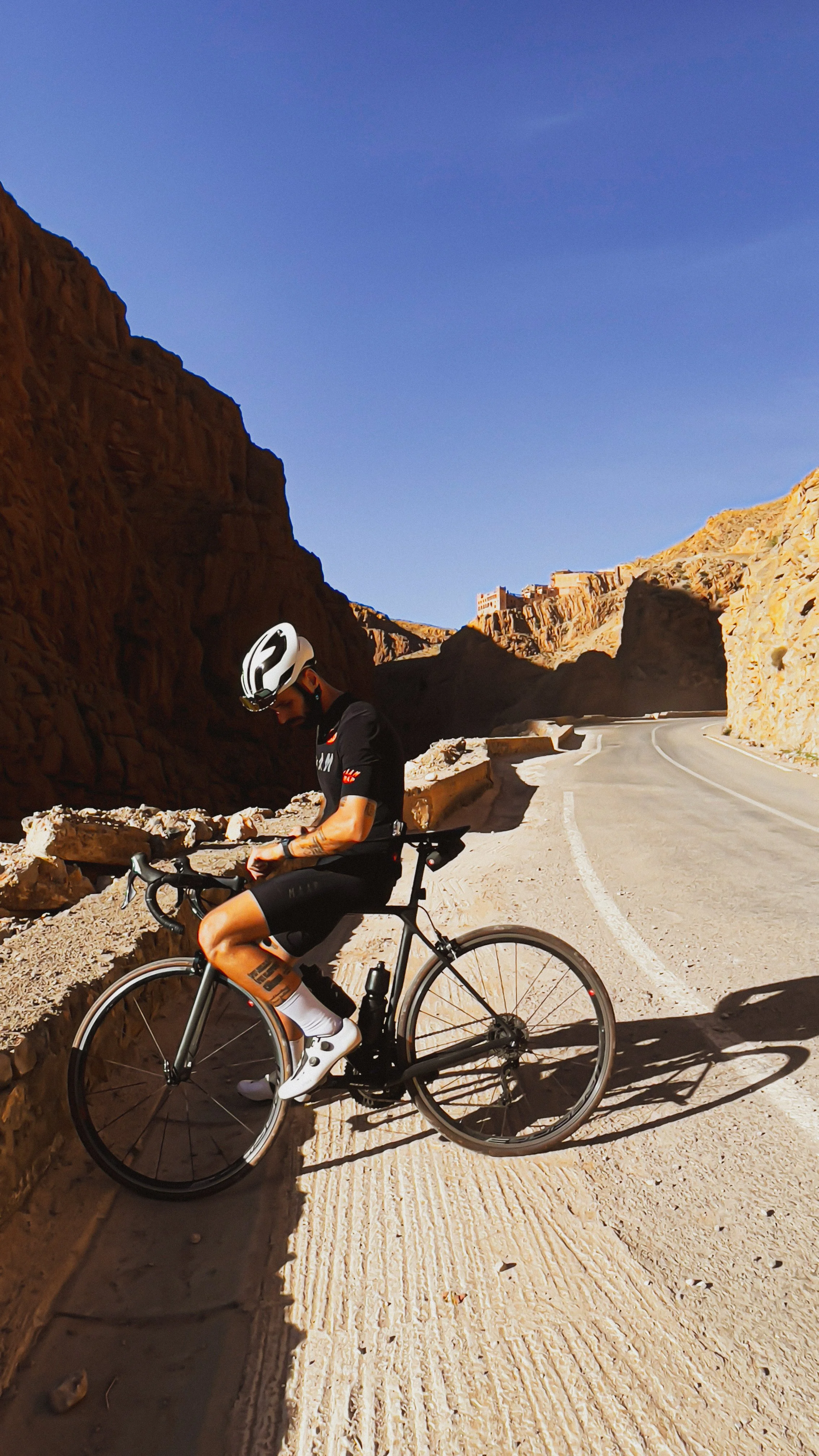 Un cycliste en tenue noire coiffé d'un casque blanc, roulant sur une route de montagne en envahir de rochers et d'une ville au sommet d'une colline, par une journée ensoleillée.
