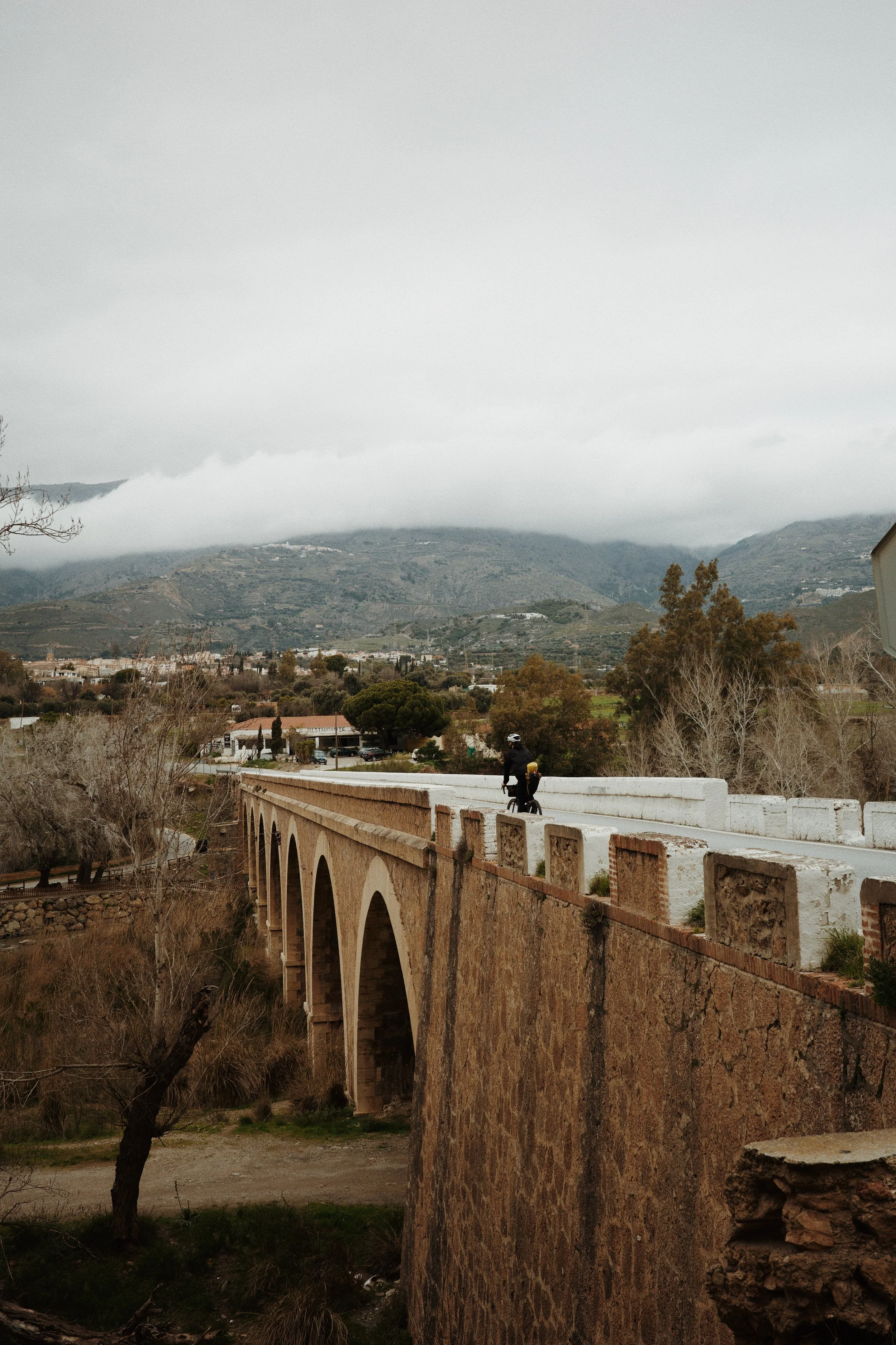 Un pont en pierre avec une personne à vélo dessus, dans un paysage de montagnes brumeuses, arbres et bâtiments en arrière-plan, ciel nuageux.