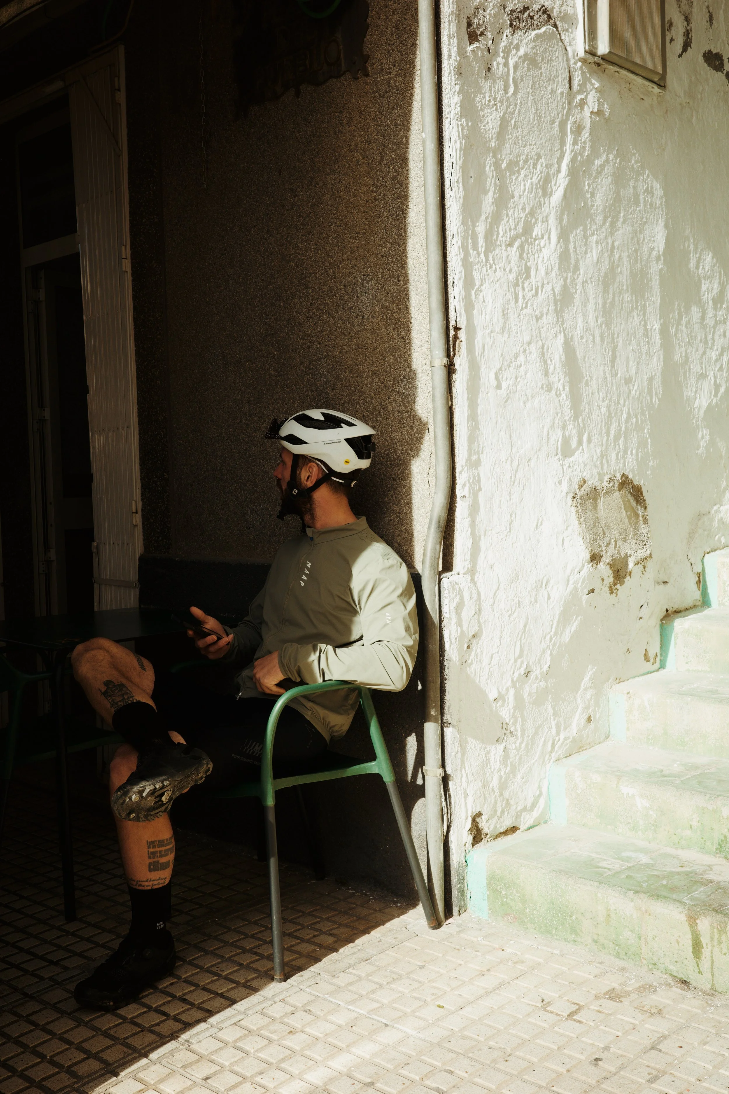 Un homme portant un casque de vélo, un sweat à capuche gris, des shorts et des chaussures de randonnée, assis sur une chaise verte en plein air, utilisant son téléphone, avec un mur en pierre blanche et une canalisation à l'arrière-plan.