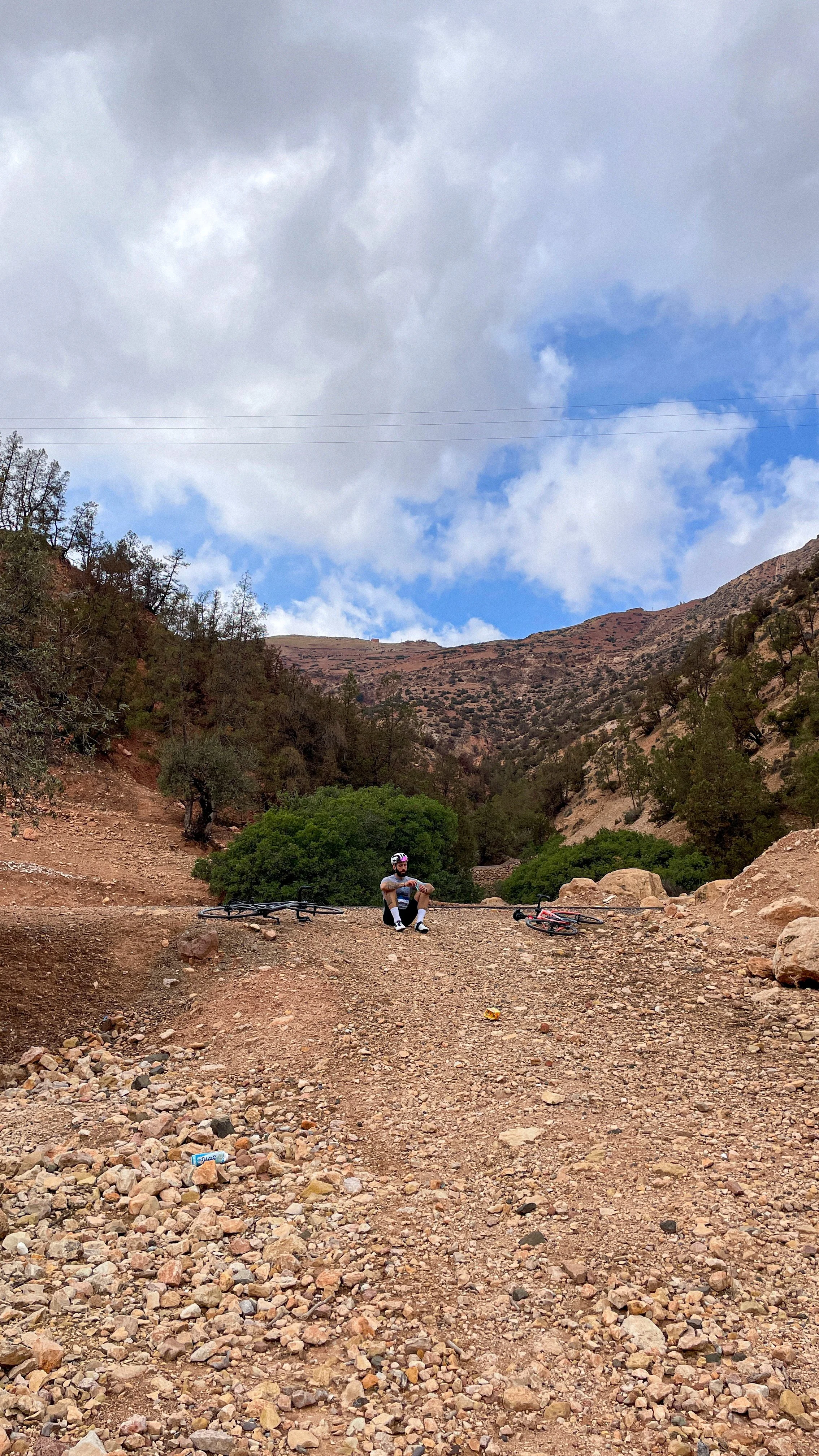 Un homme en tenue de cyclisme et casque, assis sur un terrain rocheux avec ses deux vélos couchés à côté, dans un paysage montagneux avec des arbres et un ciel nuageux.