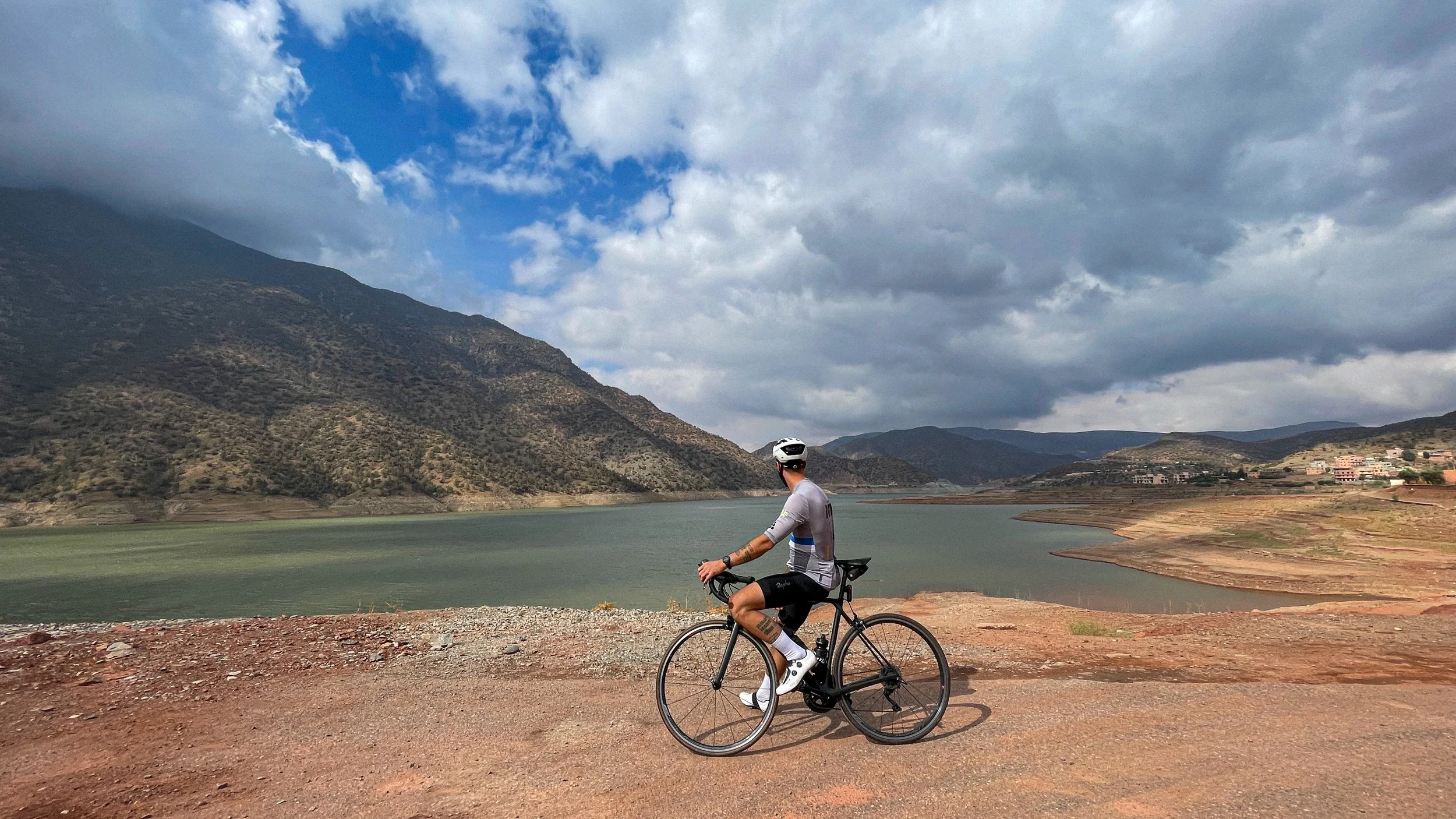 Un homme en vélo portant un casque blanc pédale près d'un lac entouré de montagnes sous un ciel nuageux.