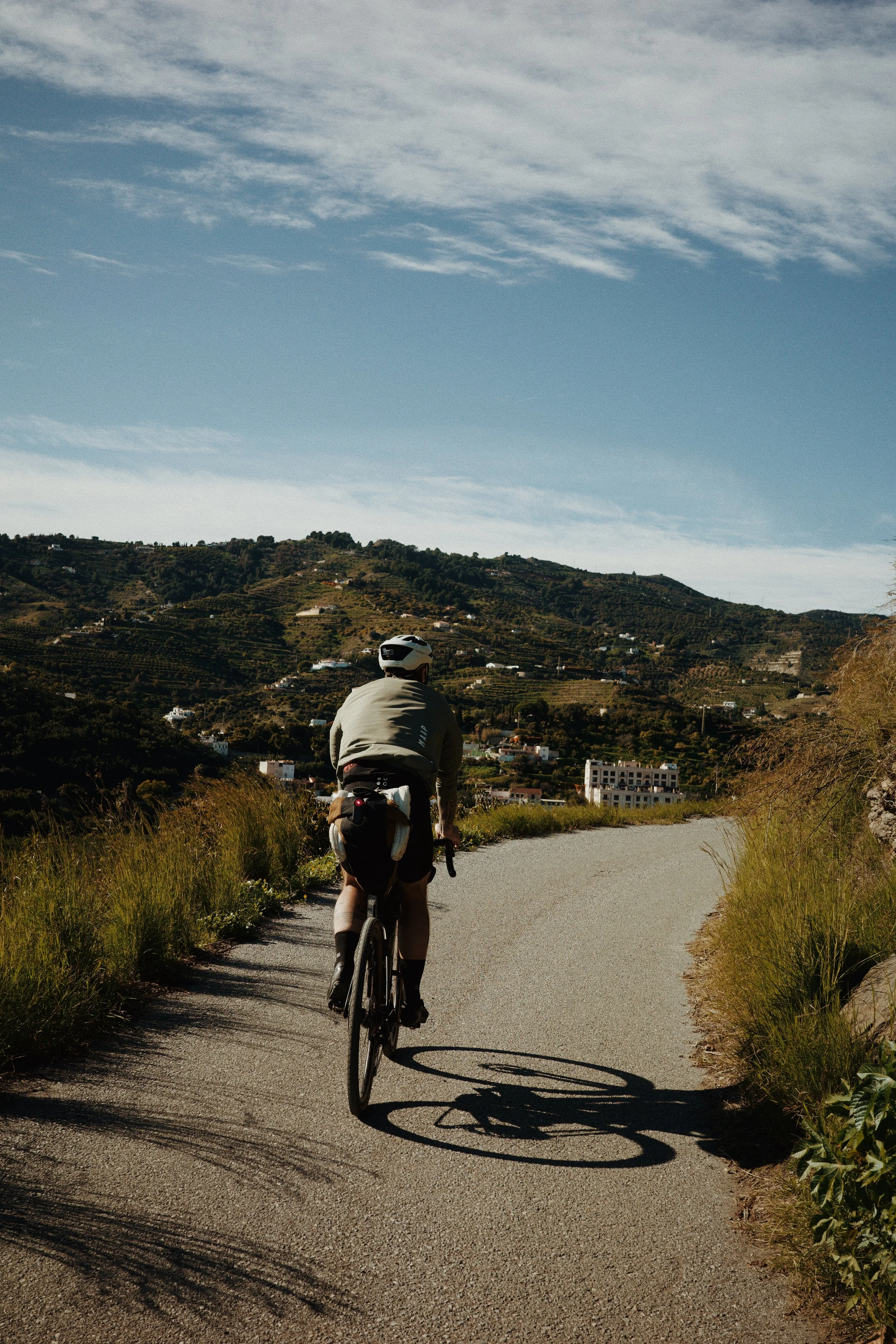 Un cycliste avec un casque et des vêtements gris, pédalant sur une route en montée dans un paysage vallonné avec des maisons à l'horizon, sous un ciel partiellement nuageux.