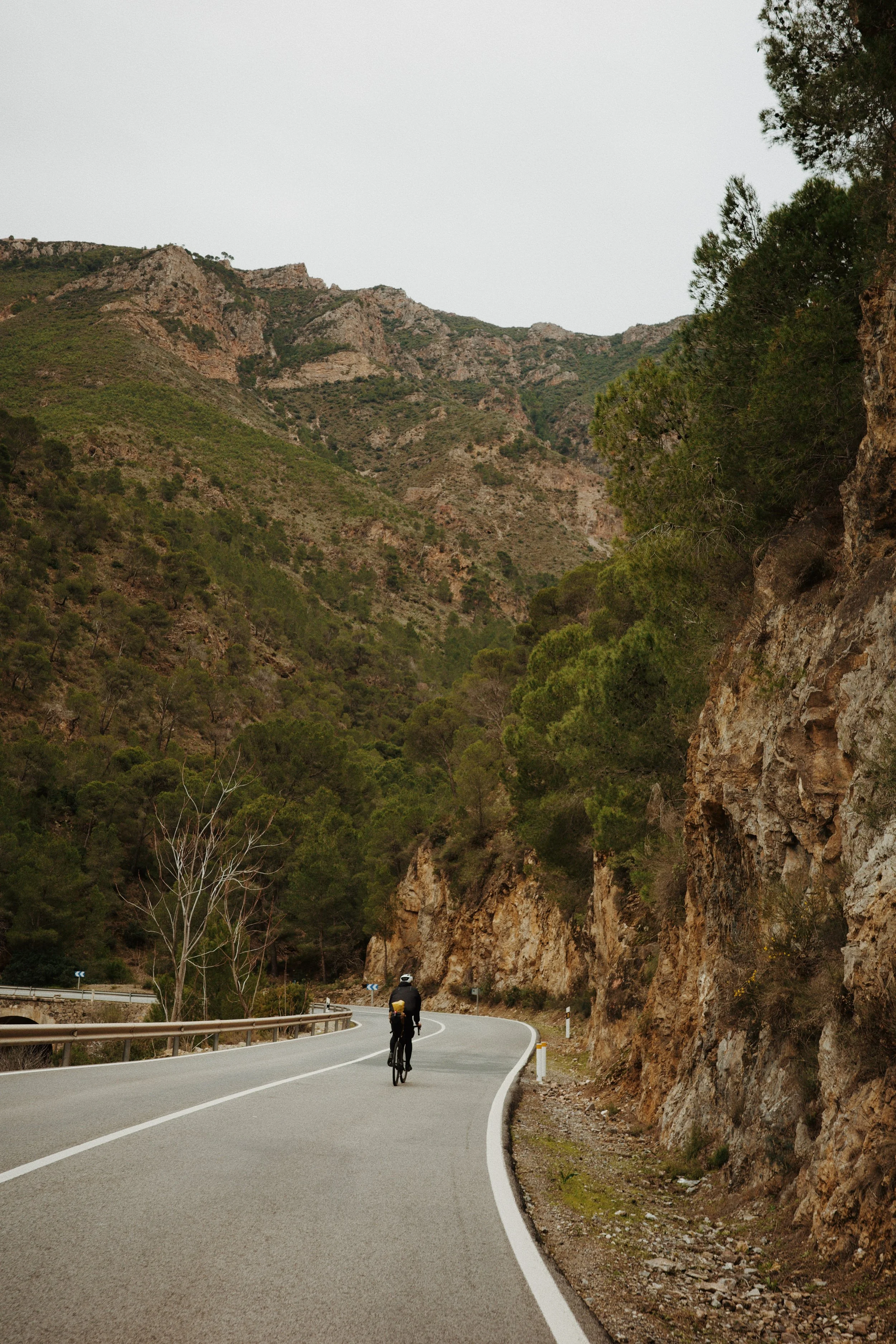 Un cycliste唯一 sur une route sinueuse entourée de montagnes rocheuses et de végétation. Le ciel est clair et légèrement nuageux.