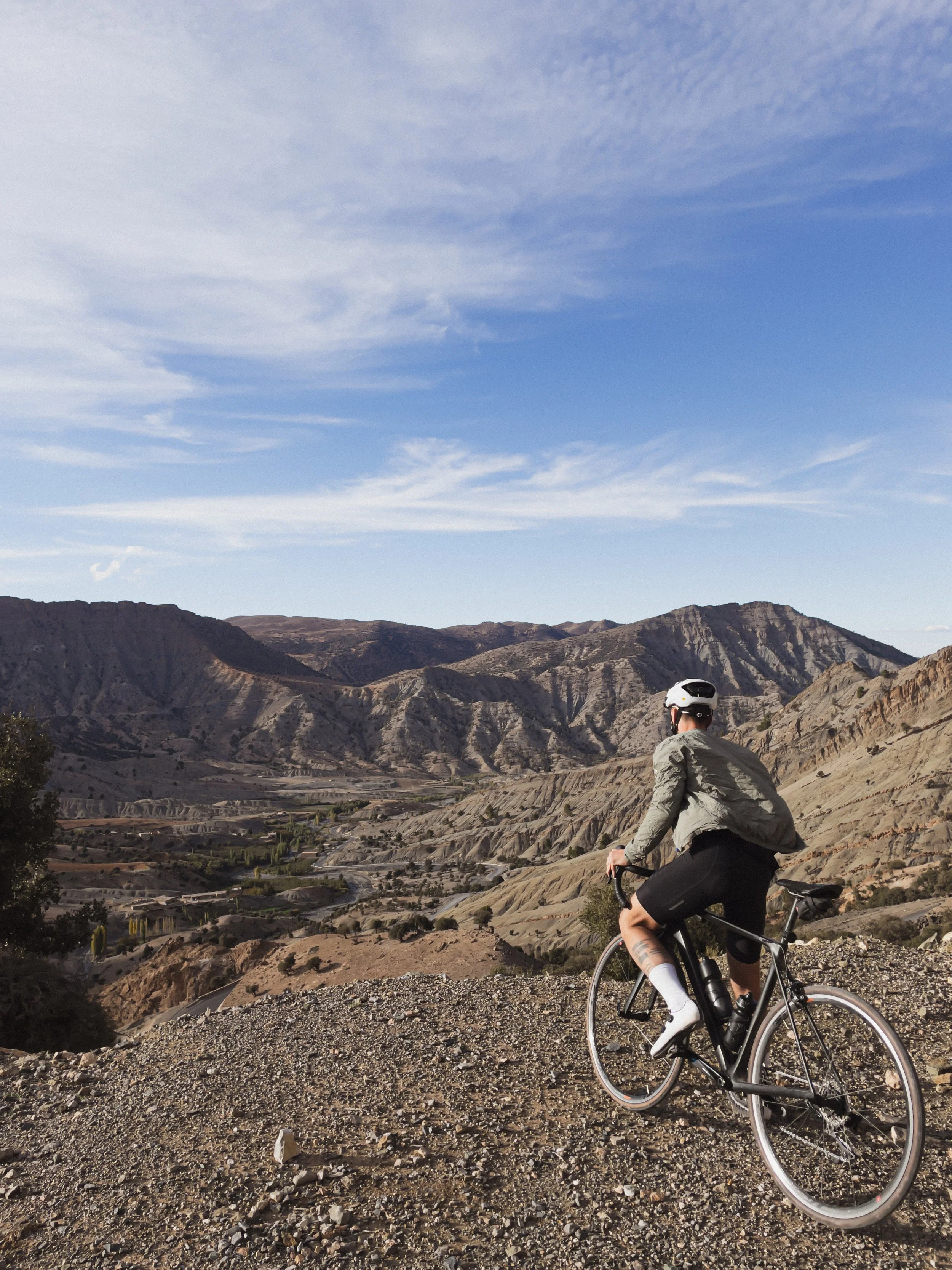 Un cycliste portant un casque traverse un paysage montagneux aride avec un ciel partiellement nuageux.