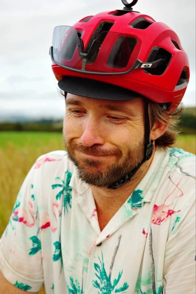 Homme avec un casque de vélo rouge et une visière, portant une chemise hawaïenne, dans un champ