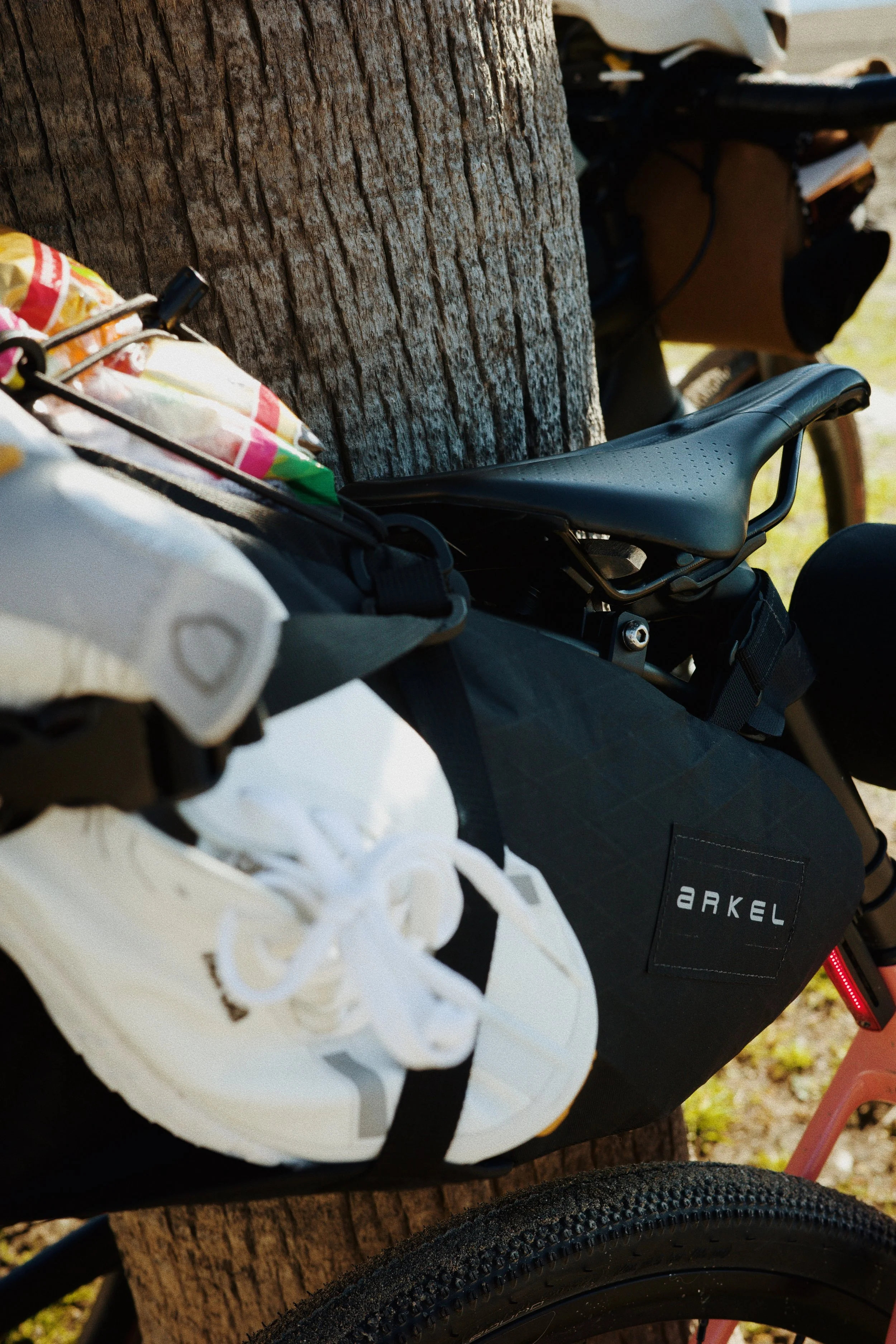 Une photo montrant un vélo avec une selle noir et un sac attaché, posé contre un arbre en extérieur, avec un casque blanc suspendu à la partie avant du vélo.