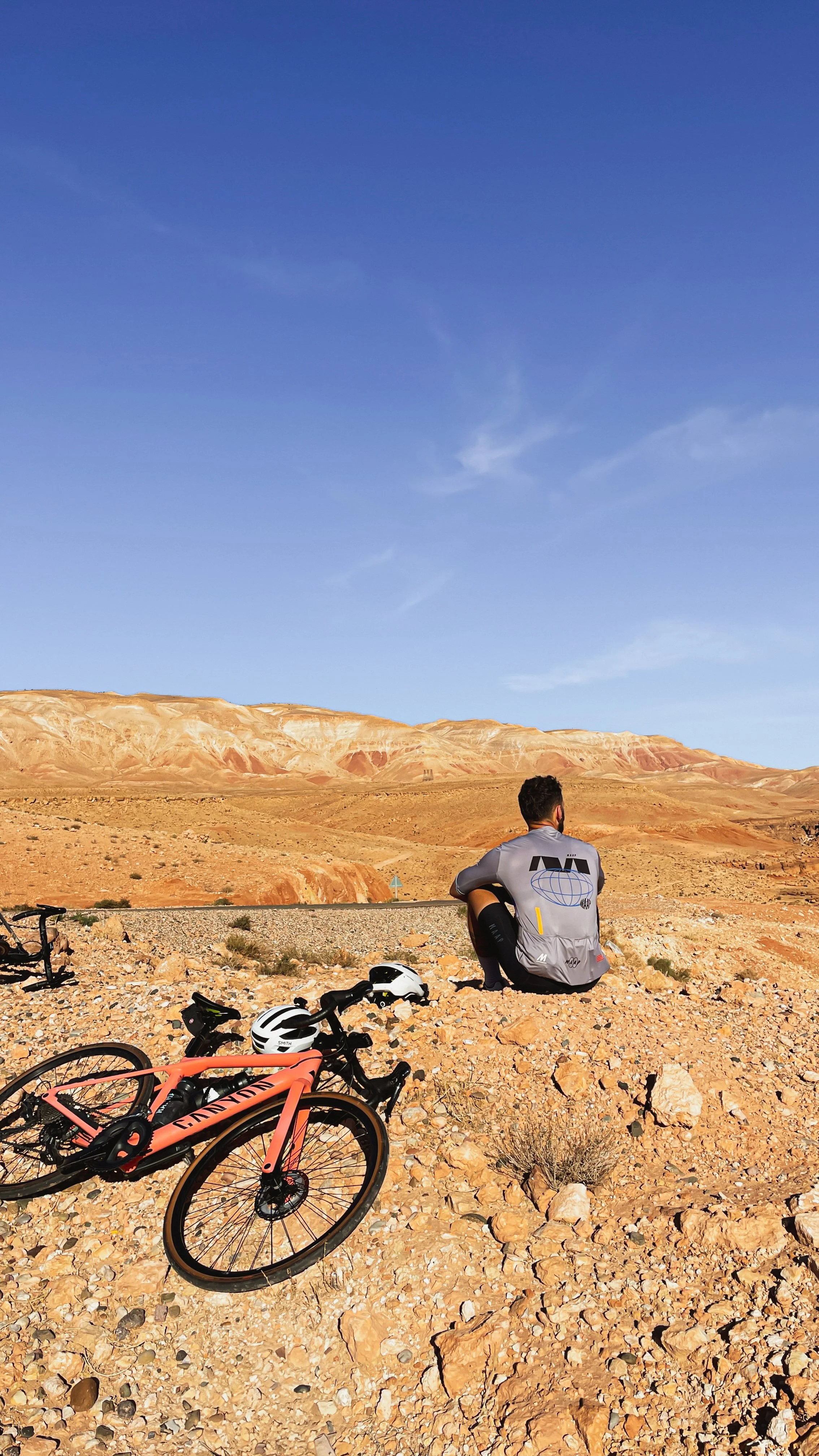 Un homme assis dans un désert rocheux avec un vélo à proximité, regardant vers un horizon de montagnes désertiques sous un ciel bleu clair.