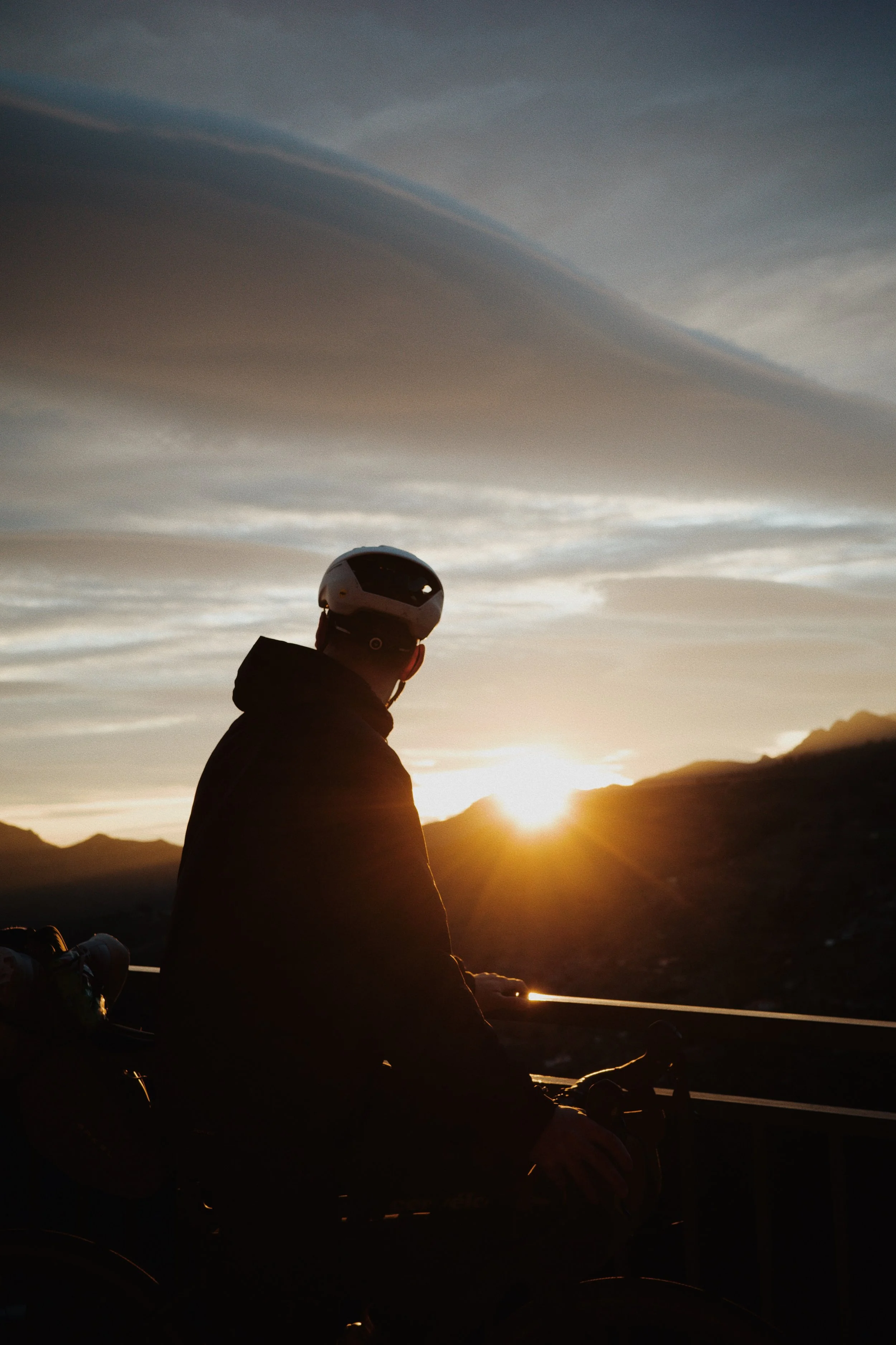 Personne avec un casque regardant un coucher de soleil, paysage montagneux en arrière-plan.