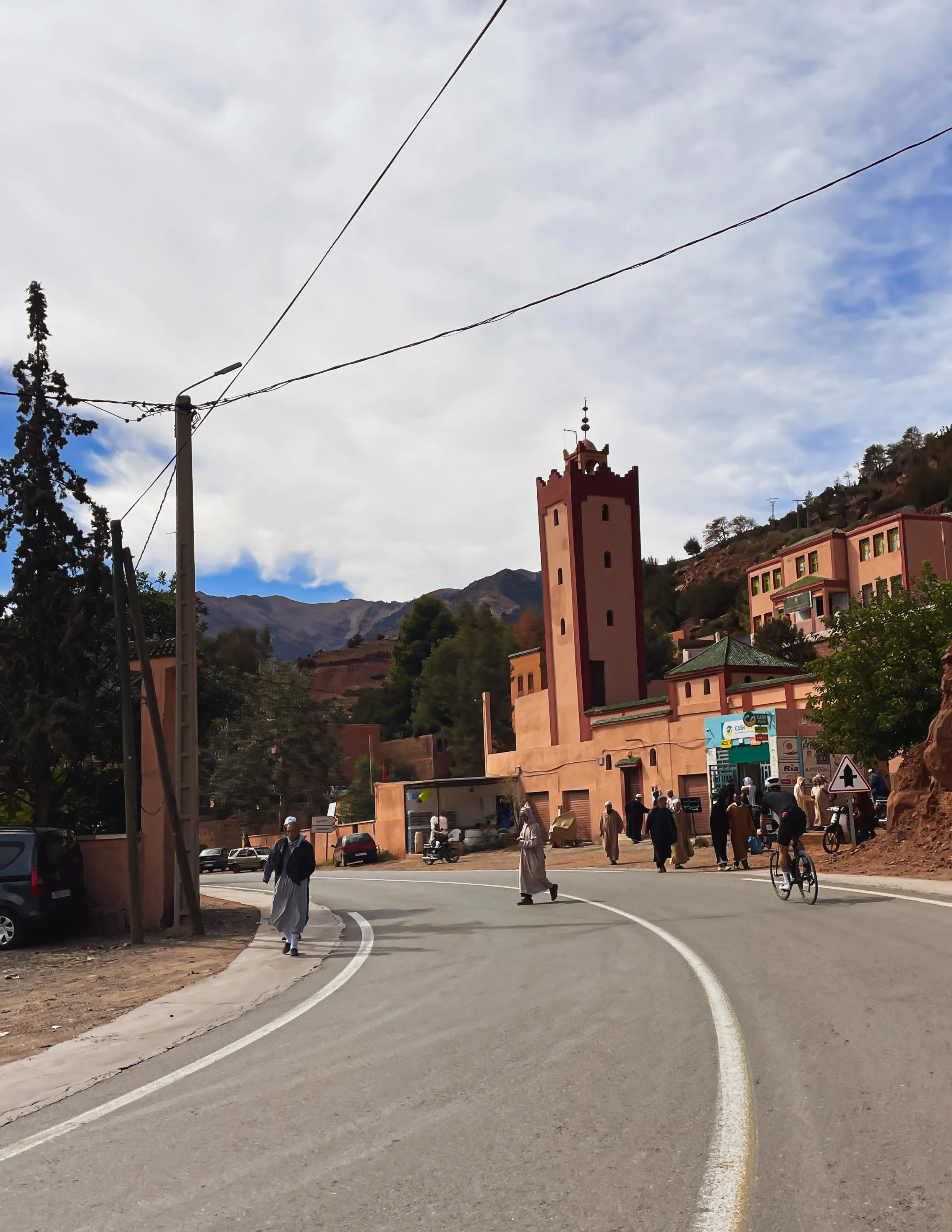 Une scène de village avec une église en arrière-plan, des personnes marchant et un cycliste, et une route sinueuse dans un environnement montagneux.