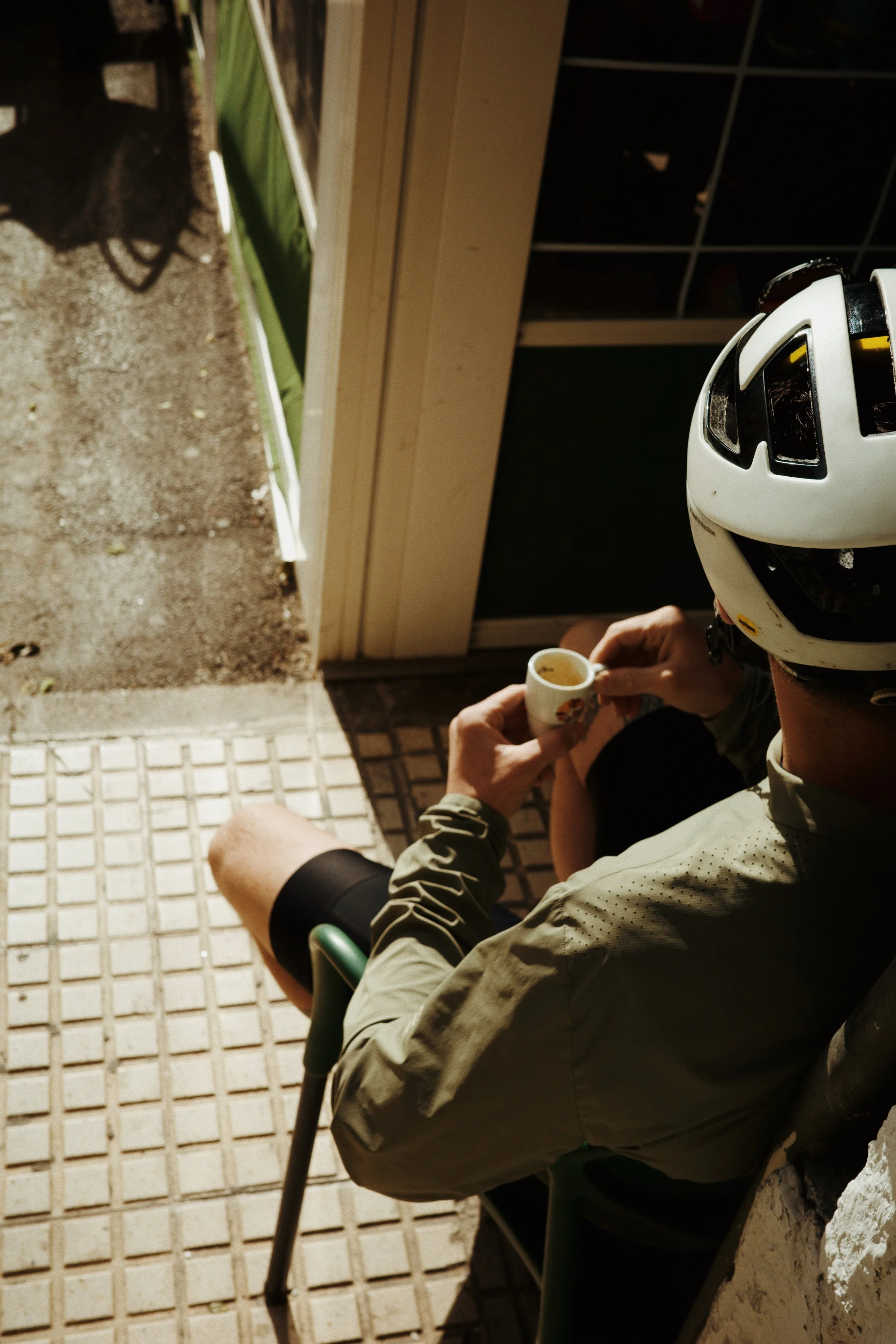 Une personne portant un casque blanc et une veste beige assise en extérieur, tenant une petite tasse, avec une maison et un sol pavé en arrière-plan.