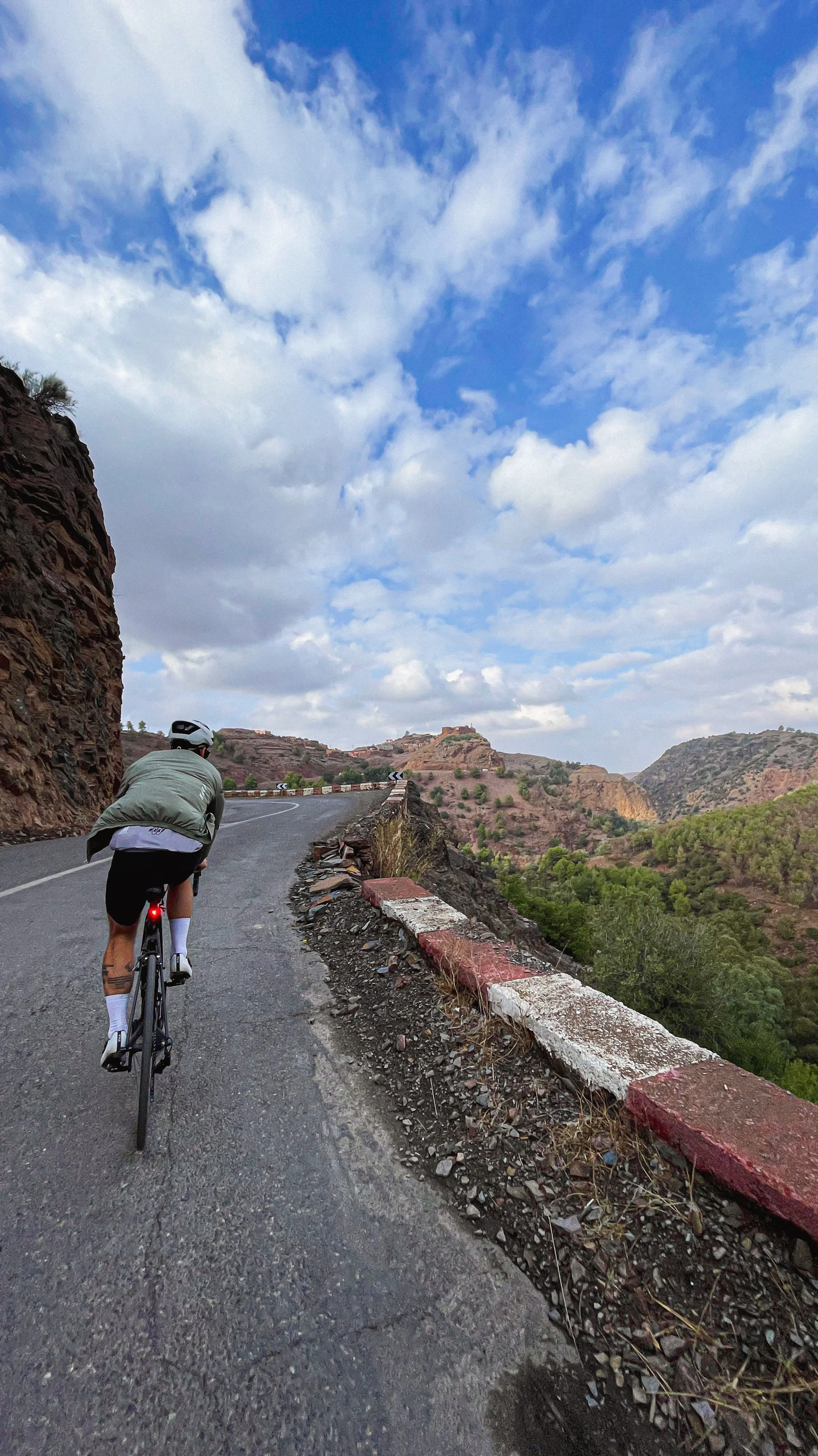 Un cycliste portant un casque et un tee-shirt porté en peignoir long, pédale sur une route montagneuse sinueuse, entourée de collines rocheuses et de végétation verte, sous un ciel partiellement nuageux.
