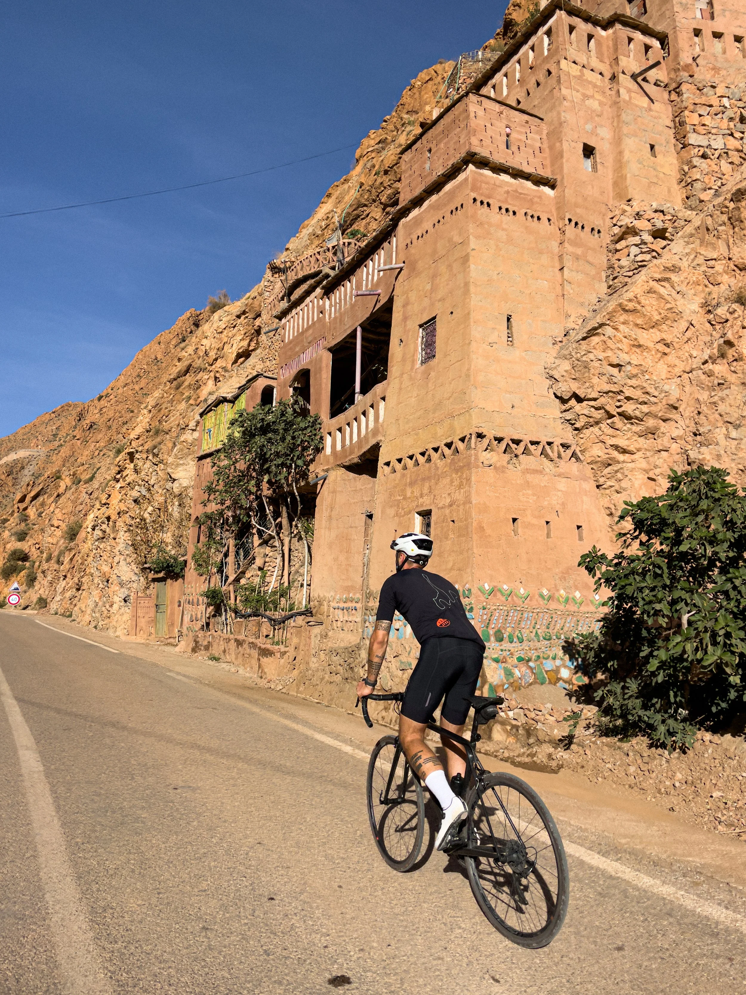 Un cycliste en tenue noire portant un casque blanc, pédale sur une route, avec une maison en adobe sur une falaise rocheuse en arrière-plan.