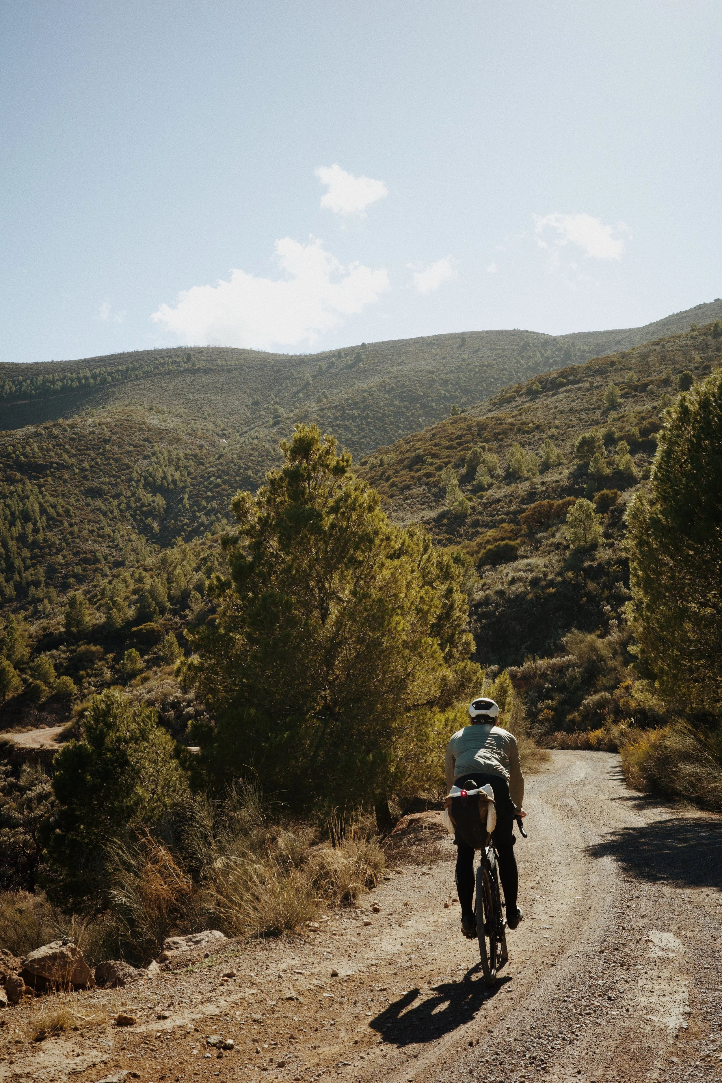 Un cycliste roulant sur un chemin de terre à travers une vallée montagneuse, entouré de végétation dense et de collines en arrière-plan, sous un ciel clair avec quelques nuages.