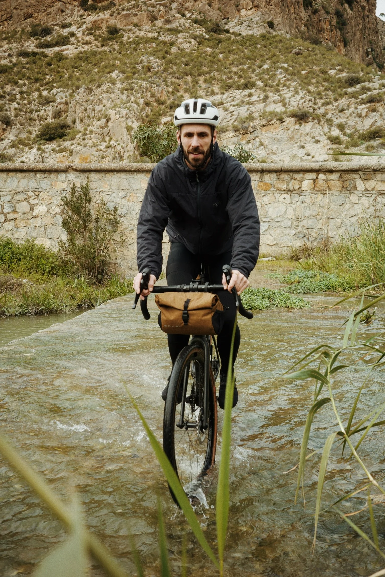 Un homme faisant du vélo à travers une rivière en montagne, portant un casque blanc et une veste noire.