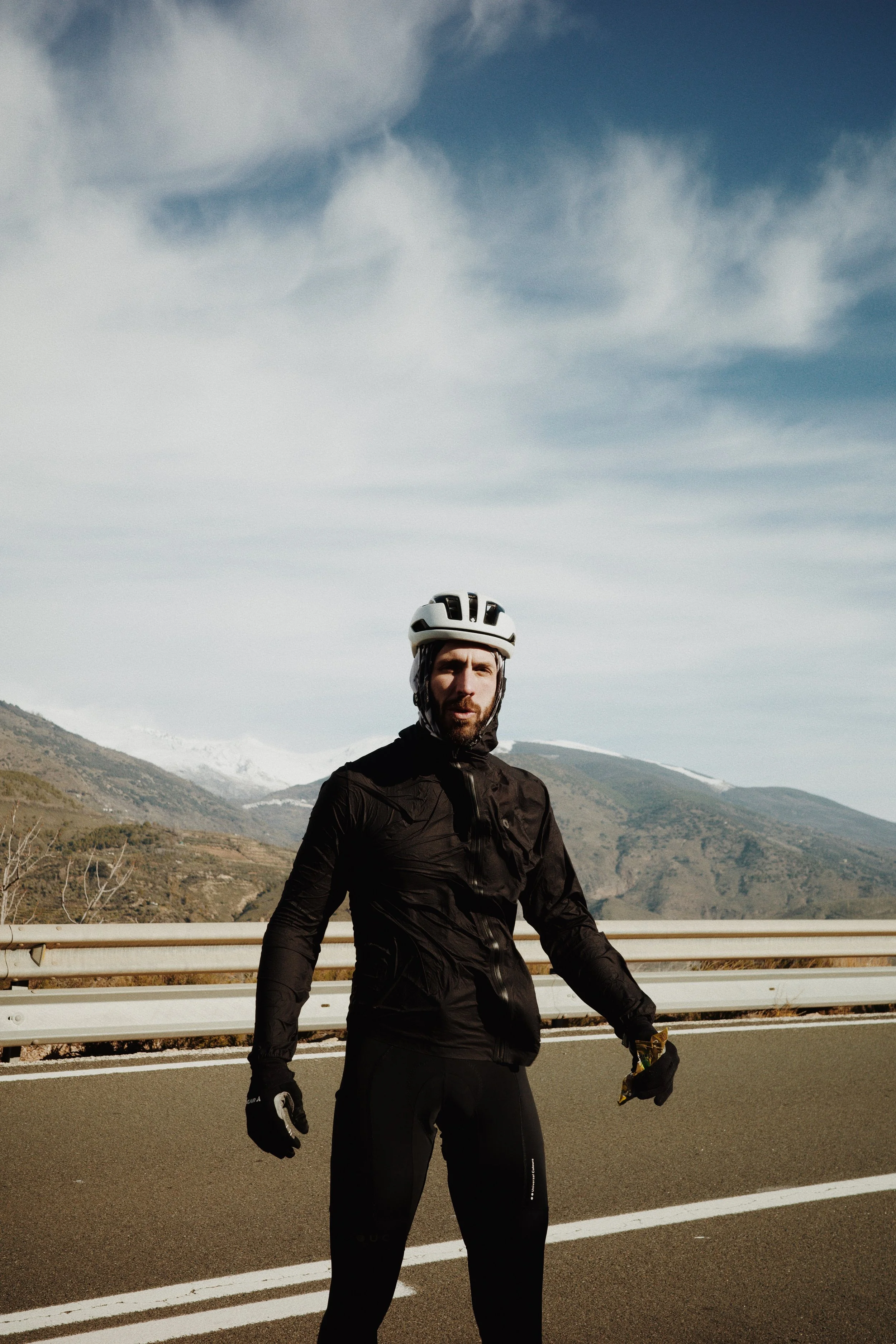 Un homme portant un casque de vélo, une veste noire et des gants, se tient sur une route avec une barrière en métal et un paysage de montagnes en arrière-plan, sous un ciel partiellement nuageux.