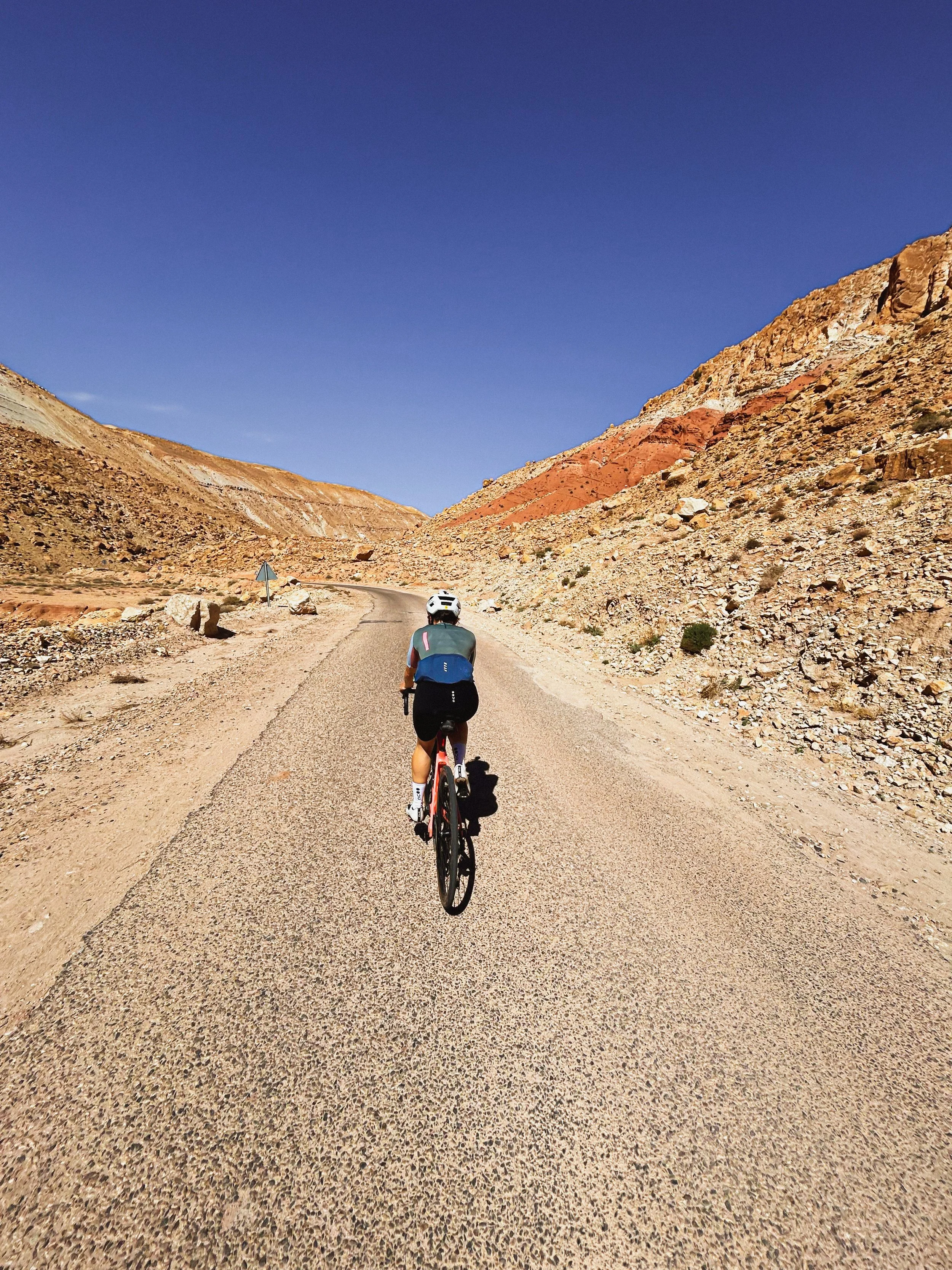 Un cycliste portant un casque pédale sur une route de montagne dans un paysage rocheux et désertique avec un ciel clair.