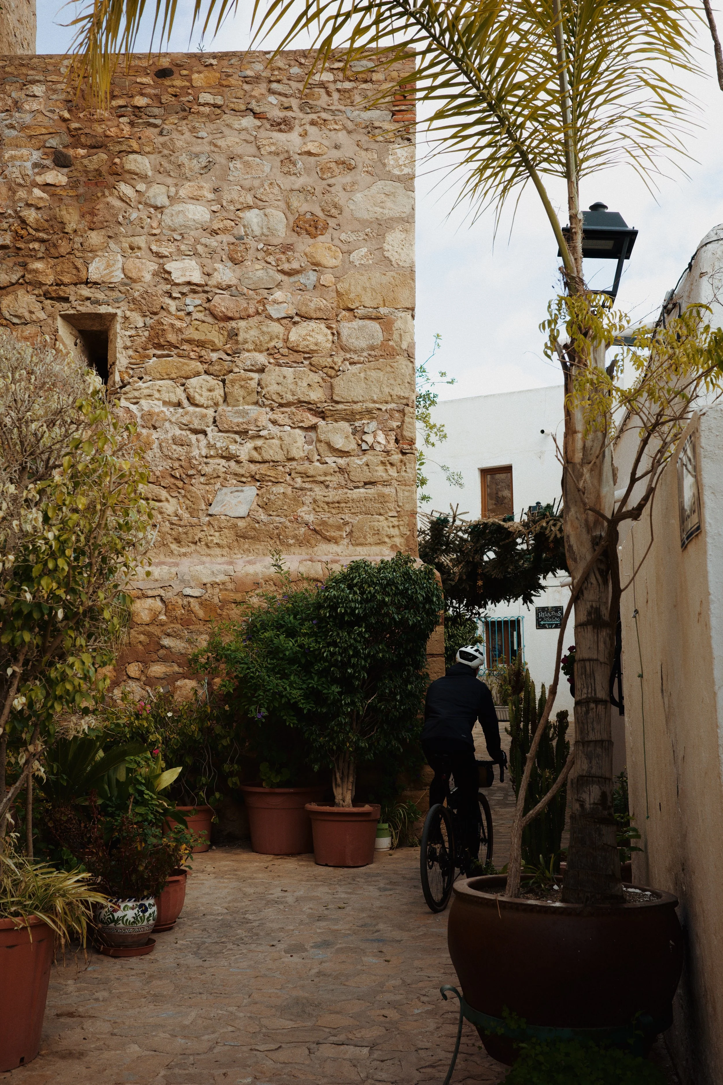 Un homme en vélo dans une cour pavée bordée de plantes en pot et un mur en pierre, avec un arbre et une lumière de lampadaire, le tout dans un style méditerranéen ou andalou.