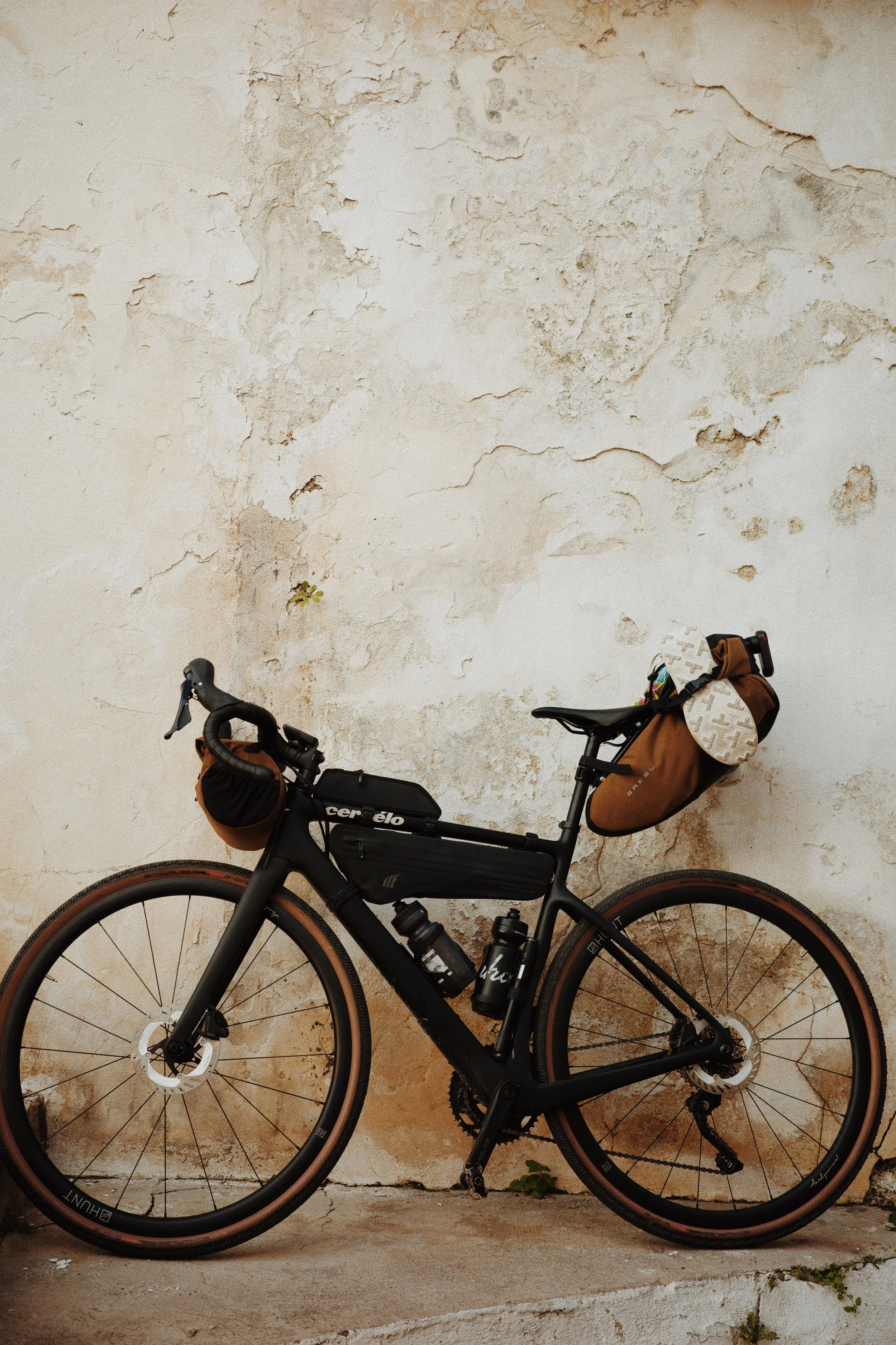 Un vélo de montagne noir avec des sacs de stockage en marron attachés à la selle et aux guidons, garé contre un mur en pierre blanche usée.