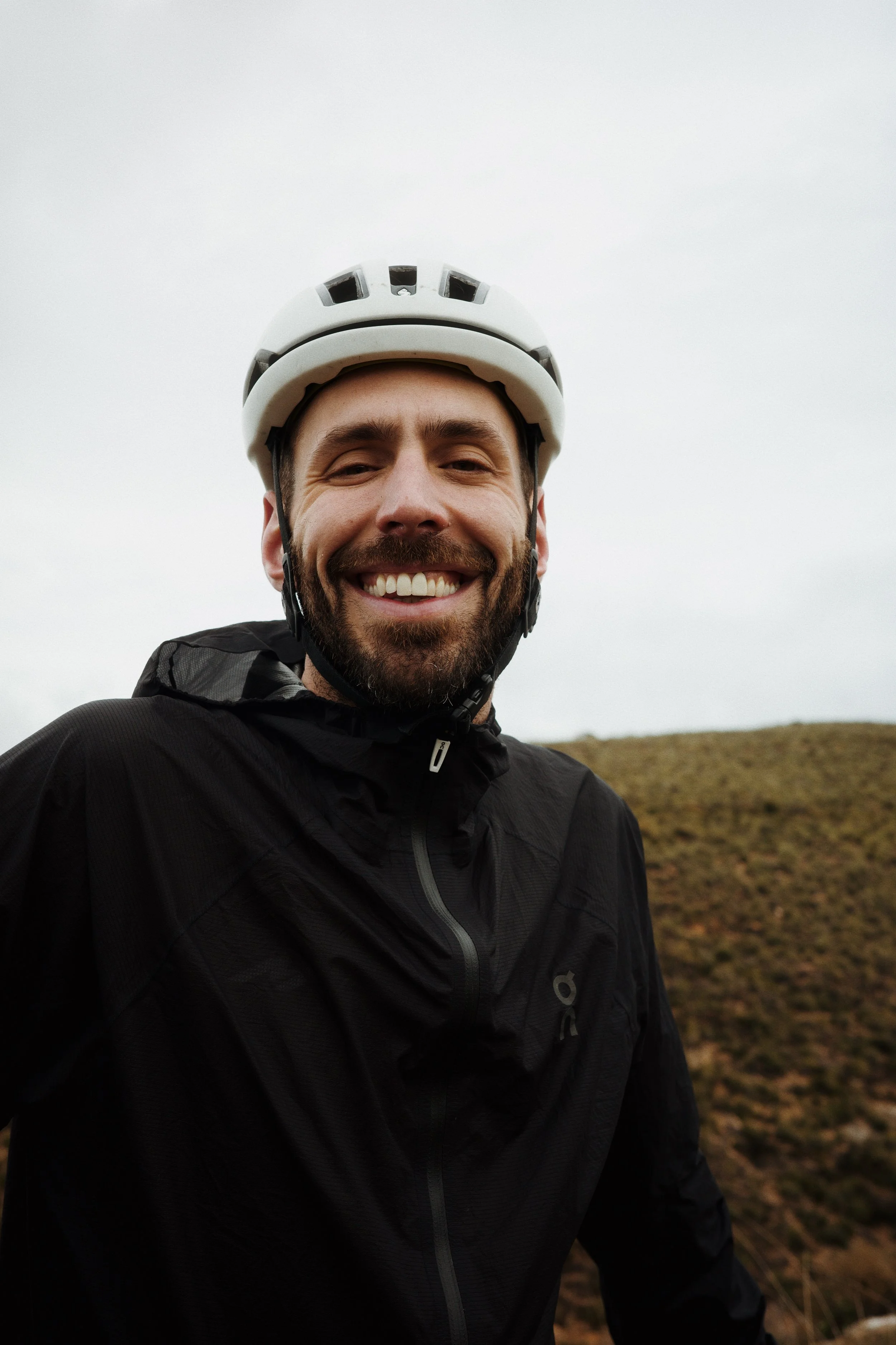 Un homme souriant portant un casque de vélo et une veste noire, en extérieur sur un terrain vallonné, ciel gris.