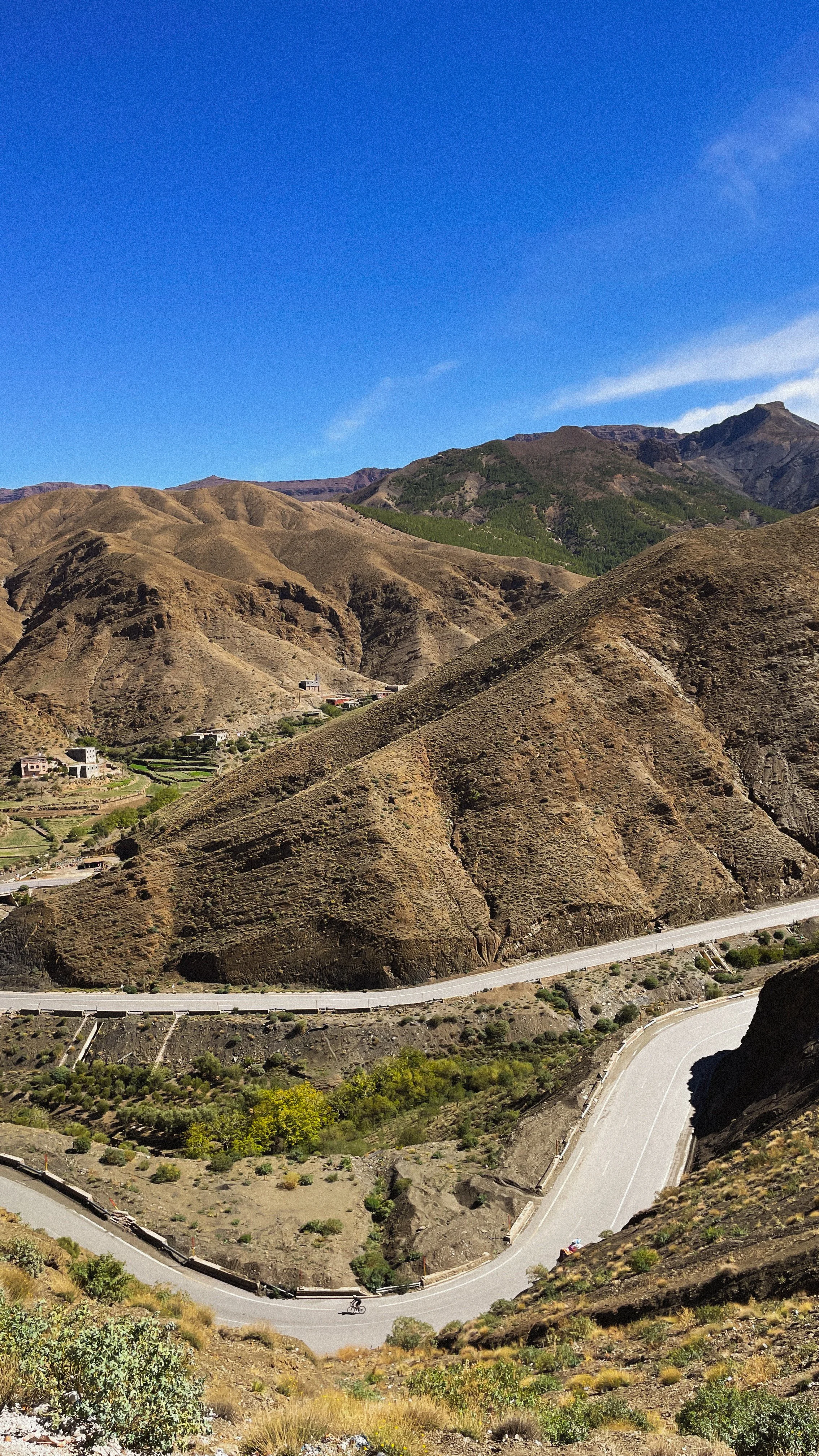 Vue panoramique de montagnes sèches avec une route sinueuse en premier plan et quelques maisons dans la vallée. Le ciel est clair et bleu.