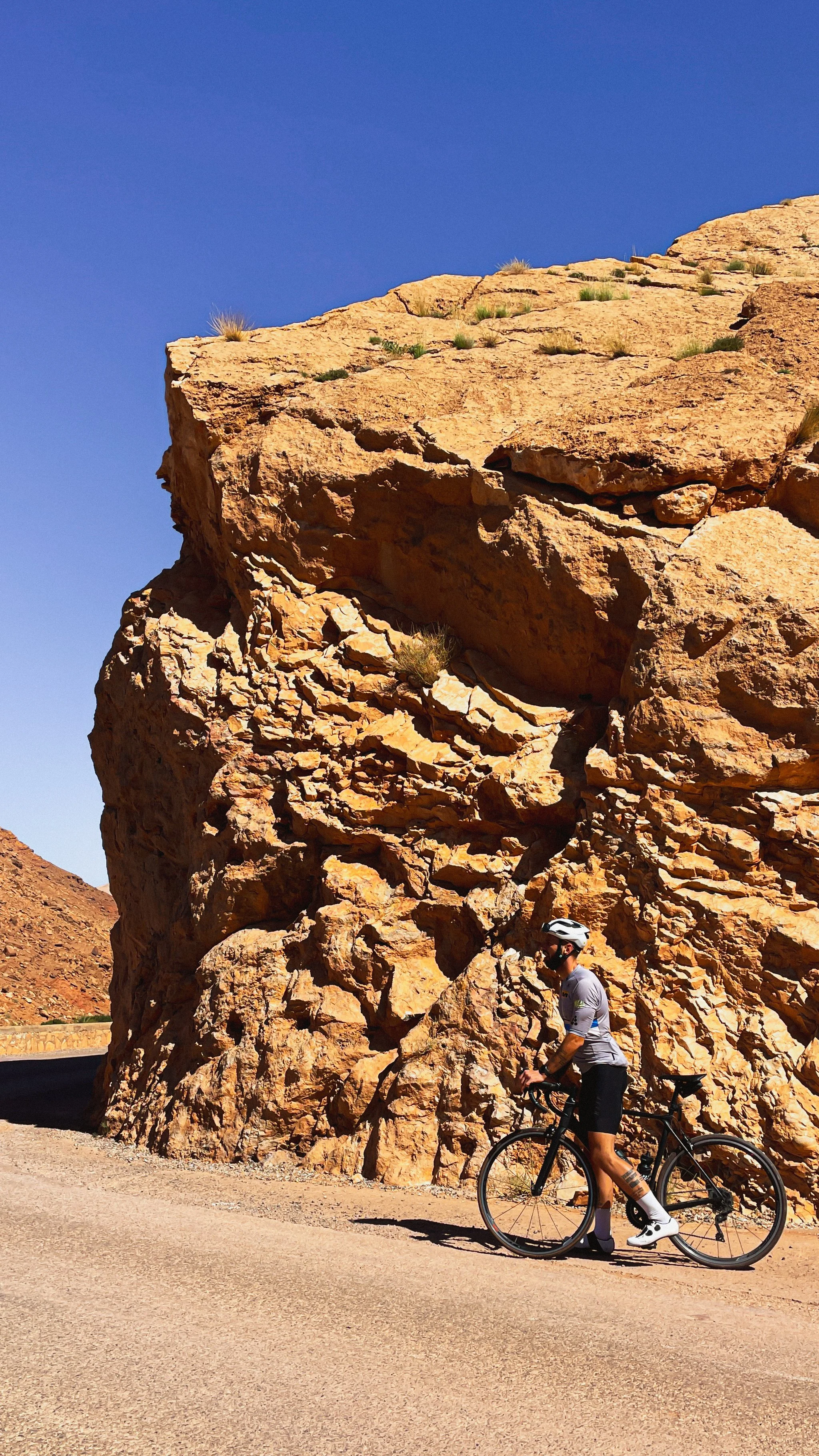 Un cycliste en tenue de sport portant un casque, qui se tient à côté de son vélo sur une route bordée de rochers rocheux dans un environnement désertique sous un ciel bleu clair.