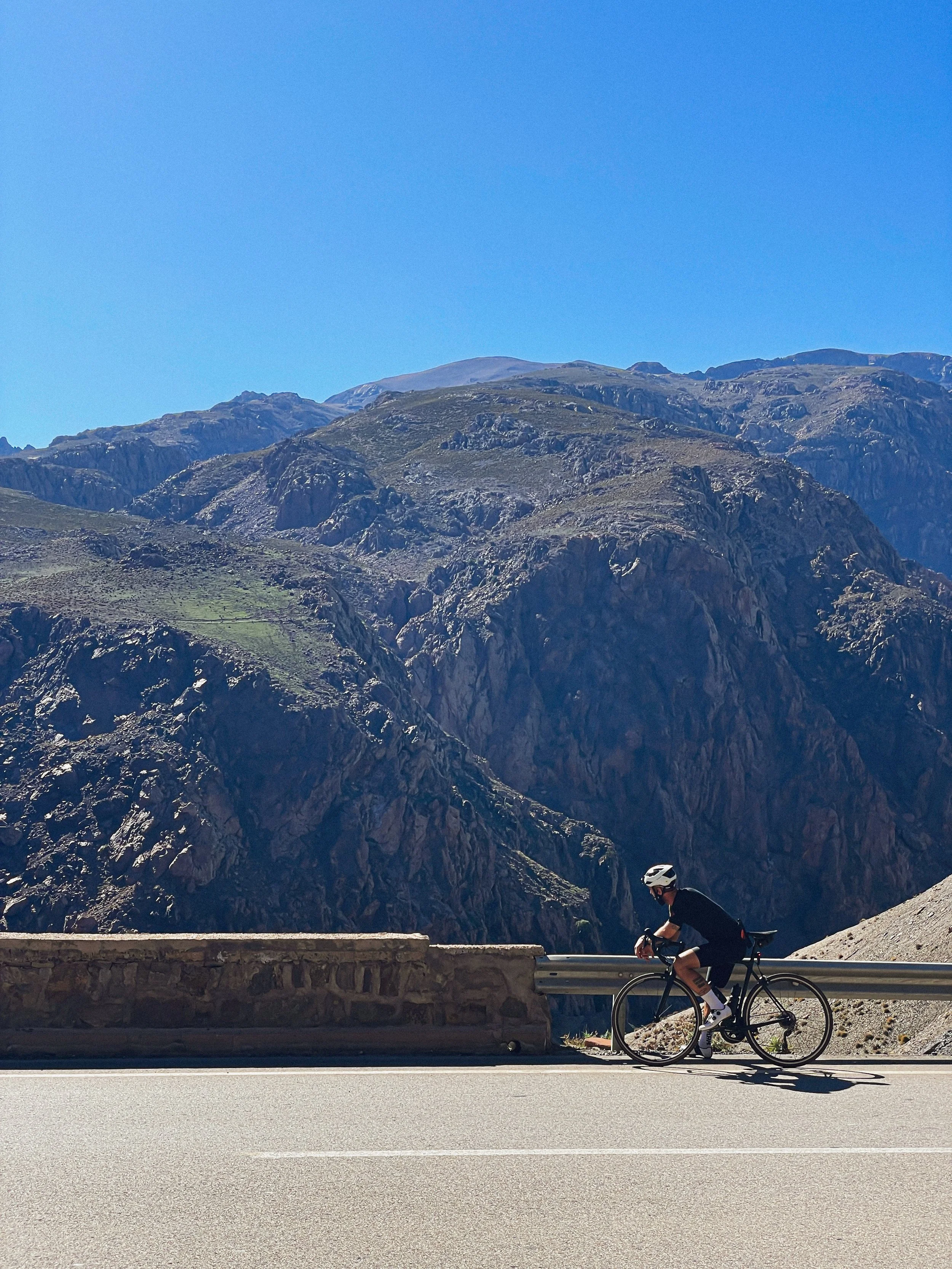 Coureur cycliste portant un casque noir et vêtement noir, pédalant sur une route en montagne avec des rochers escarpés et un ciel bleu clair.