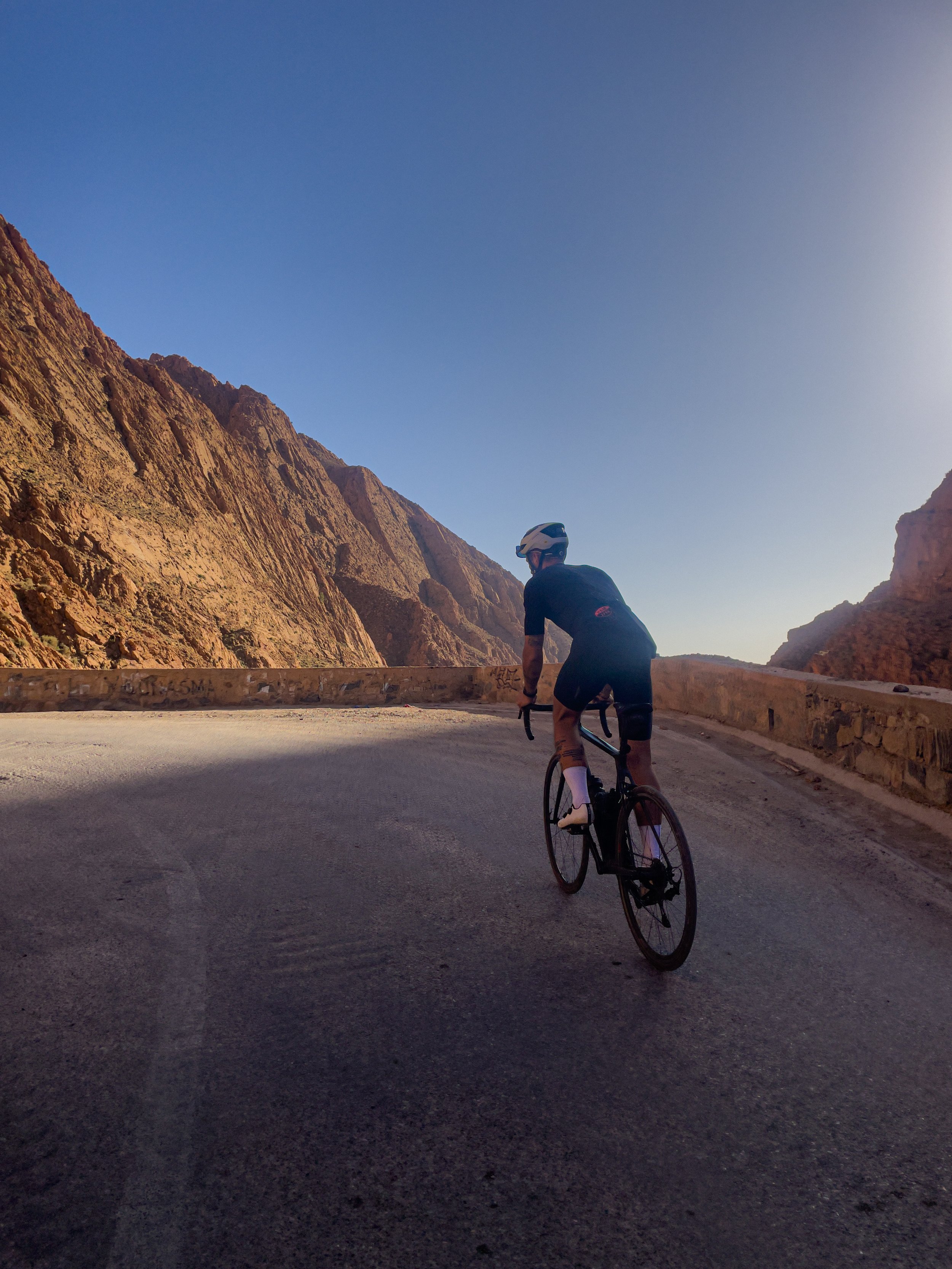 Cycliste portant un casque noir pédalant sur une route de montagne escarpée avec des parois rocheuses rocheuses sous un ciel clair.