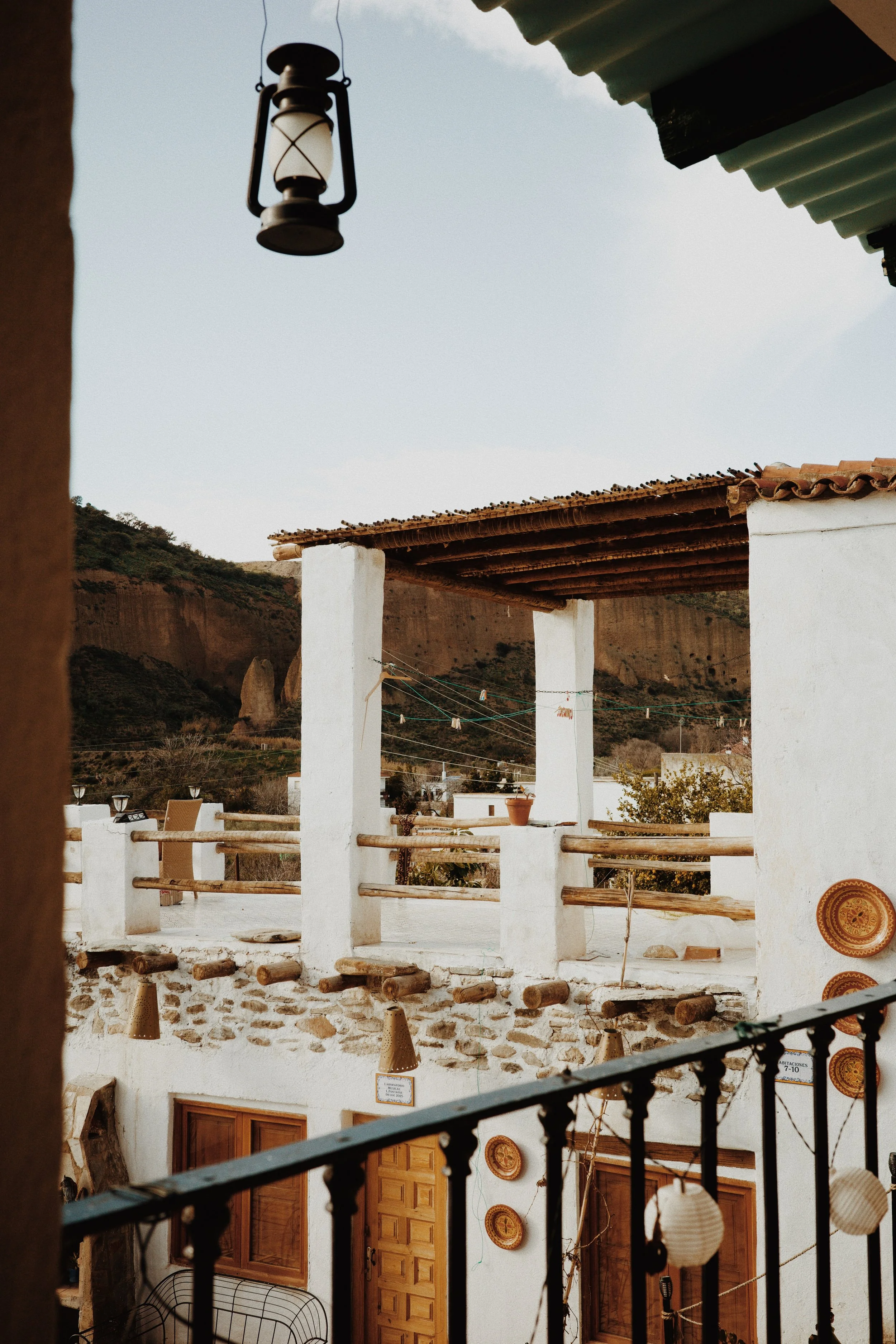 Vue d'une terrasse d'une maison de style méditerranéen avec des portes en bois et des décorations murales, entourée d'une barrière et d'une linge à linge suspendu. En arrière-plan, des collines rocheuses et un ciel clair.