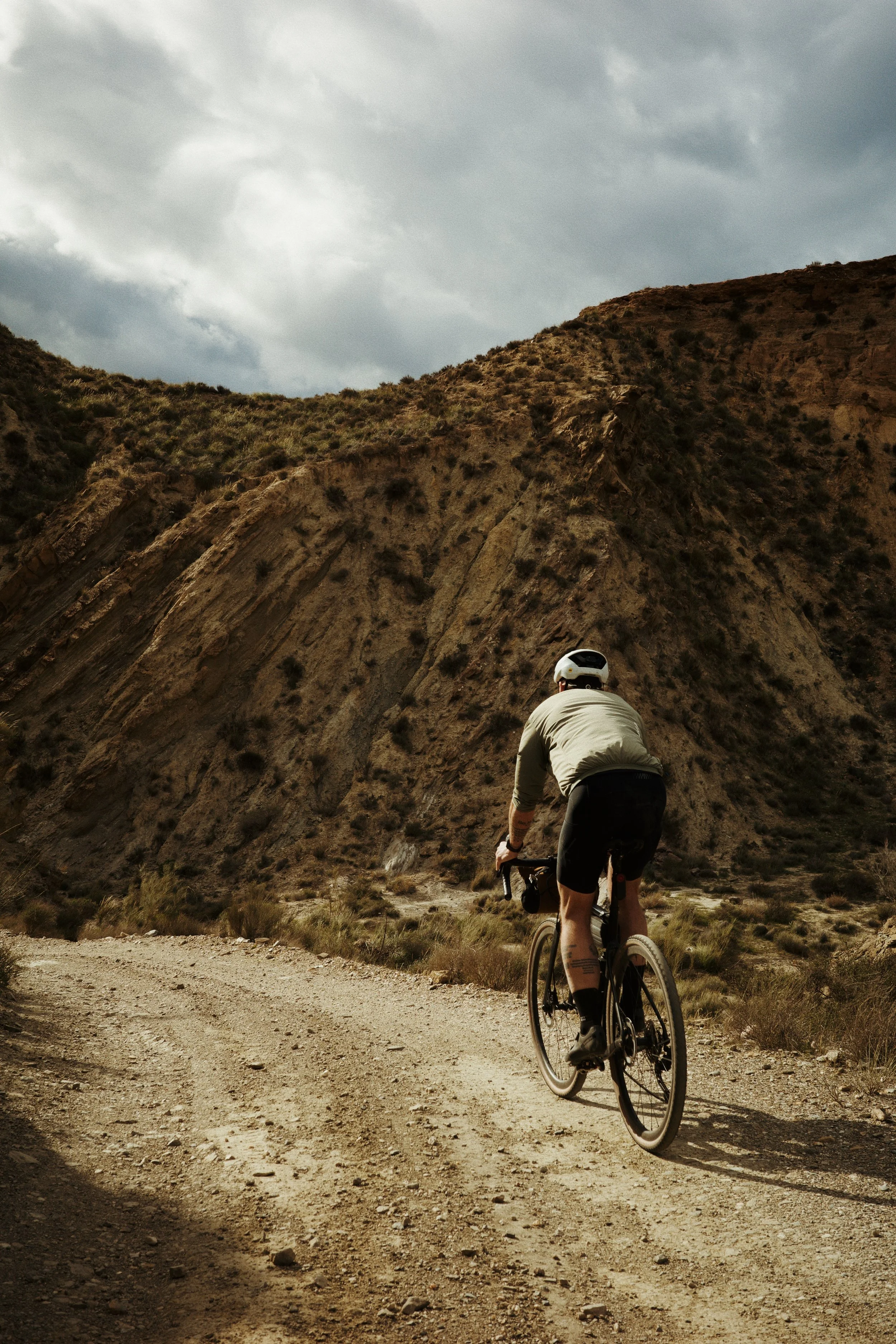 Un cycliste habillé d'un t-shirt gris, portant un casque blanc, pédalant sur une route de terre dans un paysage désertique avec des collines rocheuses et un ciel nuageux.