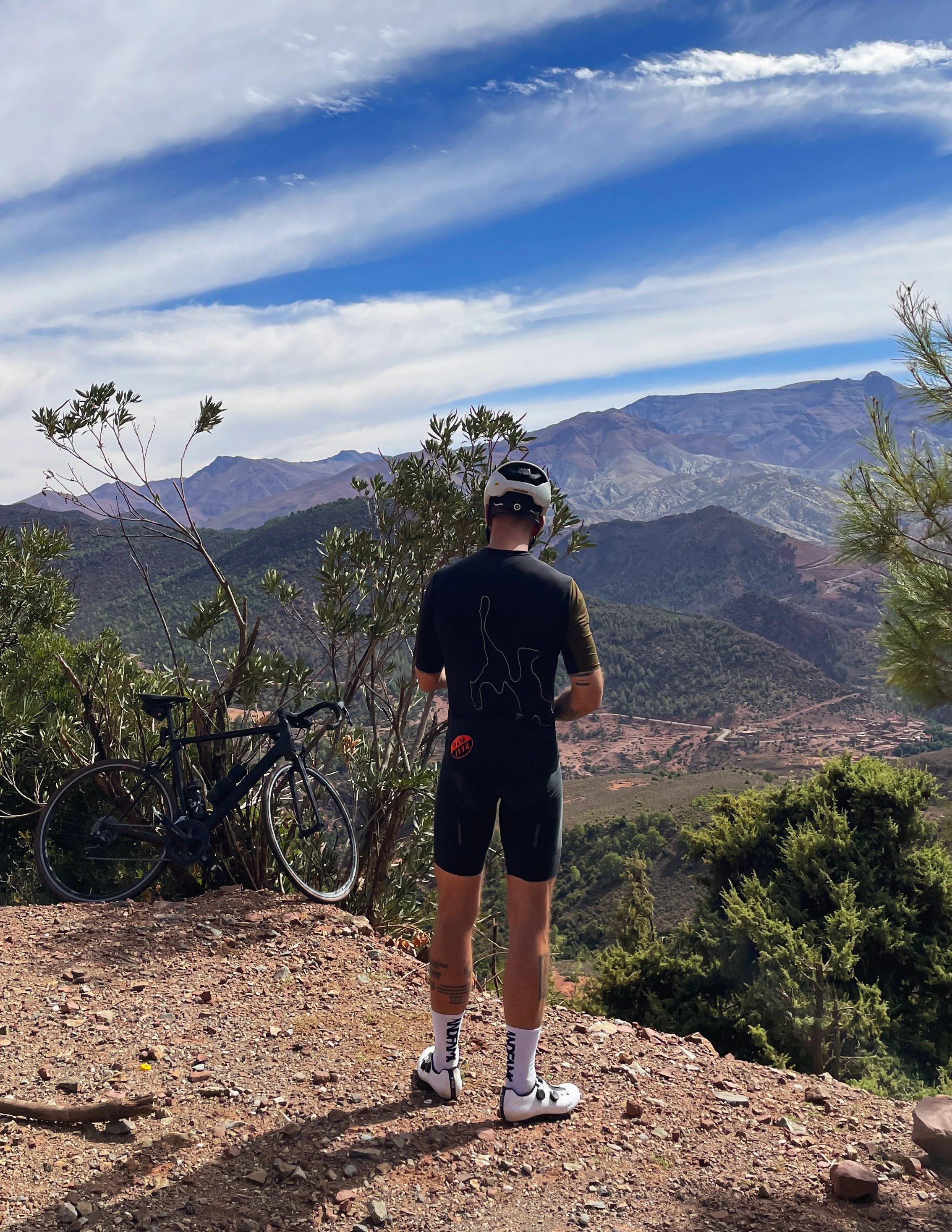 Un homme en tenue de cycliste portant un casque, vu de dos, regarde un paysage de montagnes avec peu de végétation, sous un ciel partiellement nuageux.