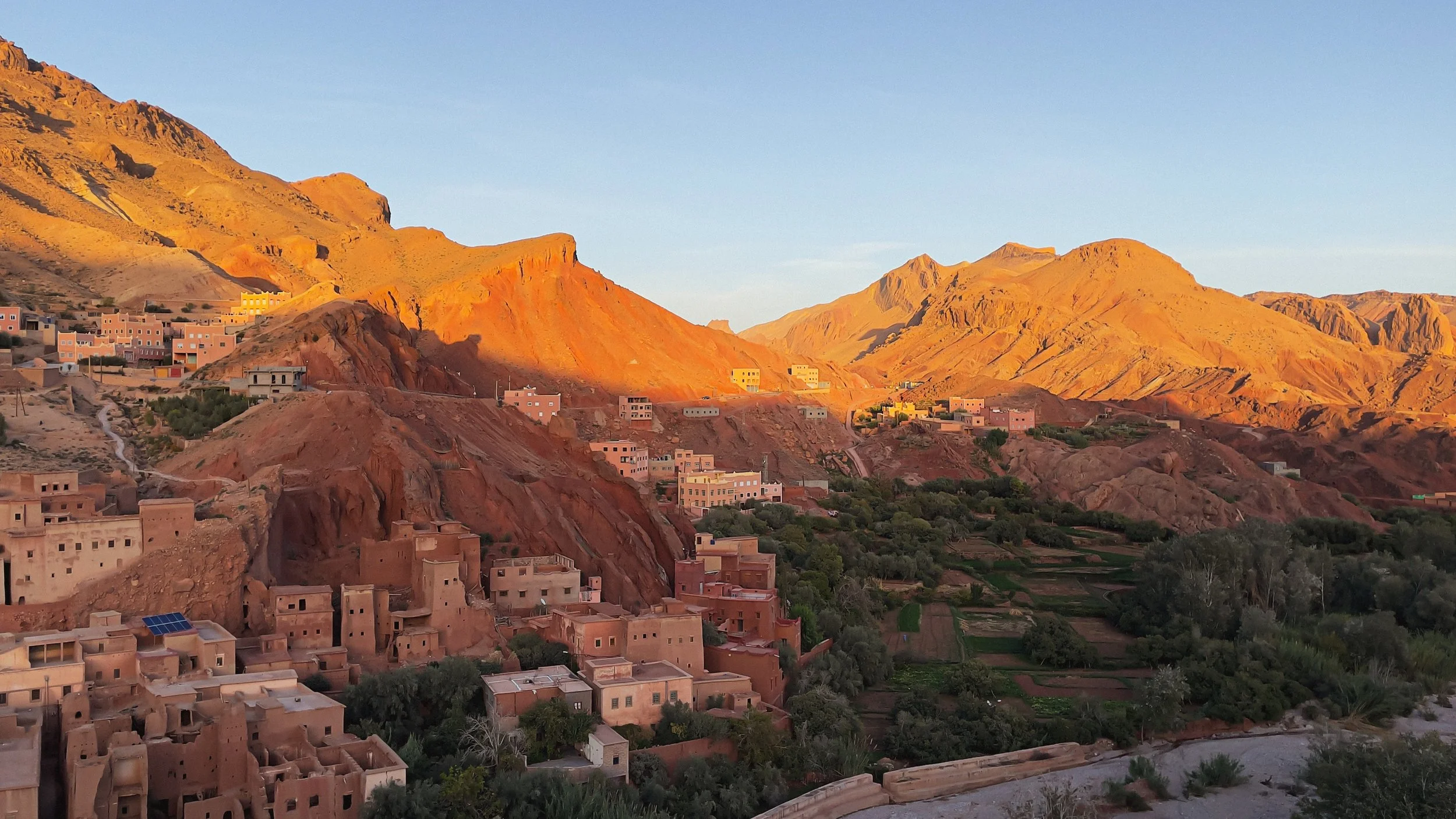 Paysage montagneux avec des maisons en adobe aux couleurs chaudes à l'avant-plan et des montagnes en arrière-plan sous un ciel clair.