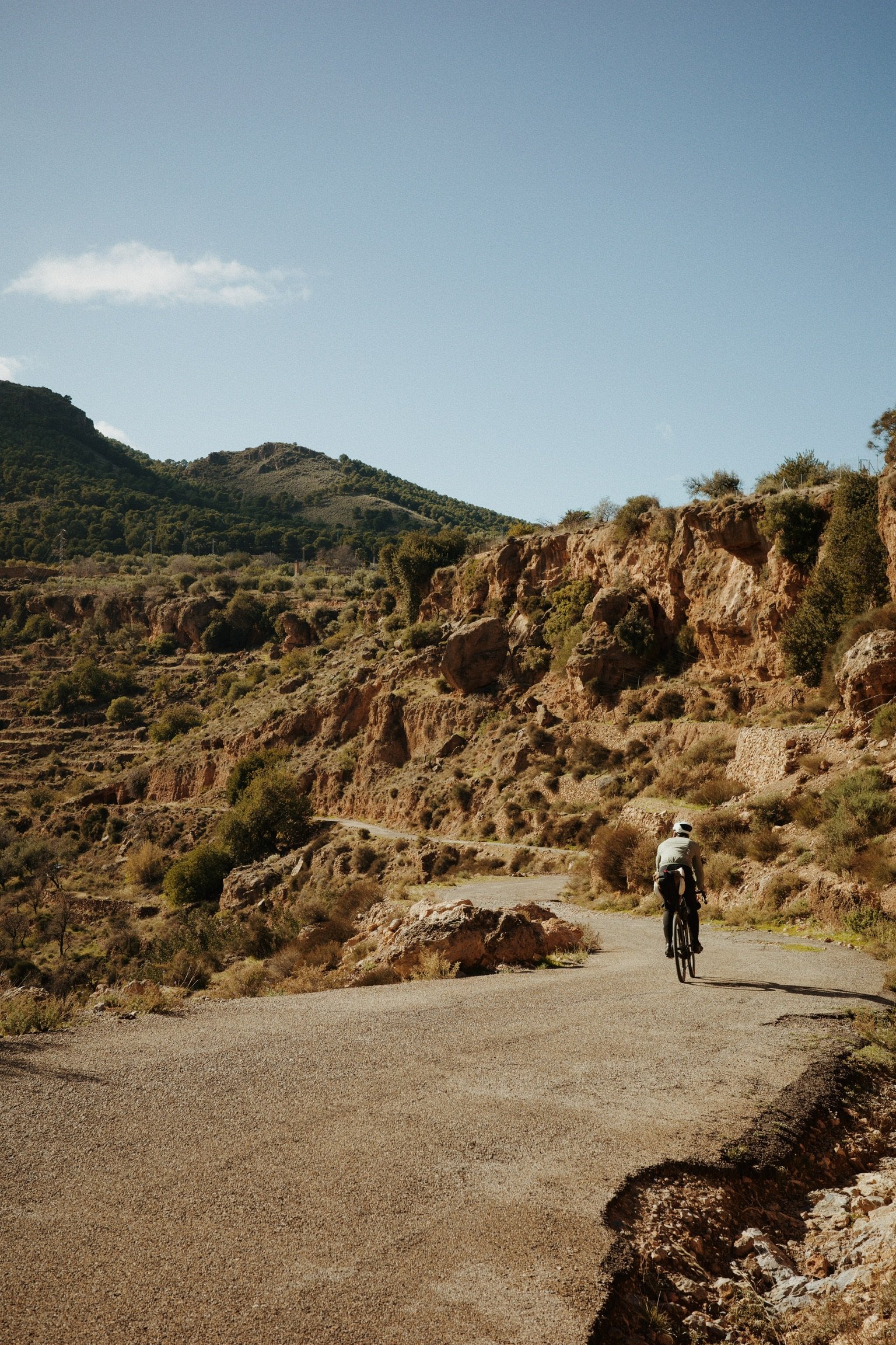 Un cycliste pédale sur une route de montagne entourée de collines rocheuses et de végétation sèche, sous un ciel bleu clair.