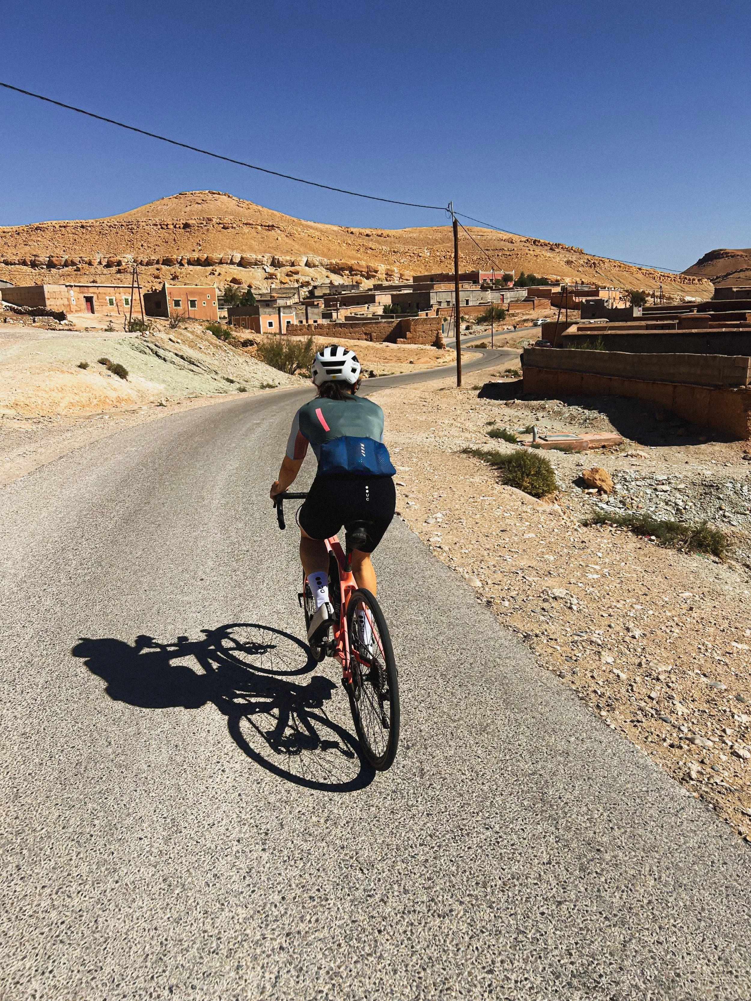 Une personne faisant du vélo dans un paysage désertique avec des collines, des maisons en adobe, et un ciel clair.