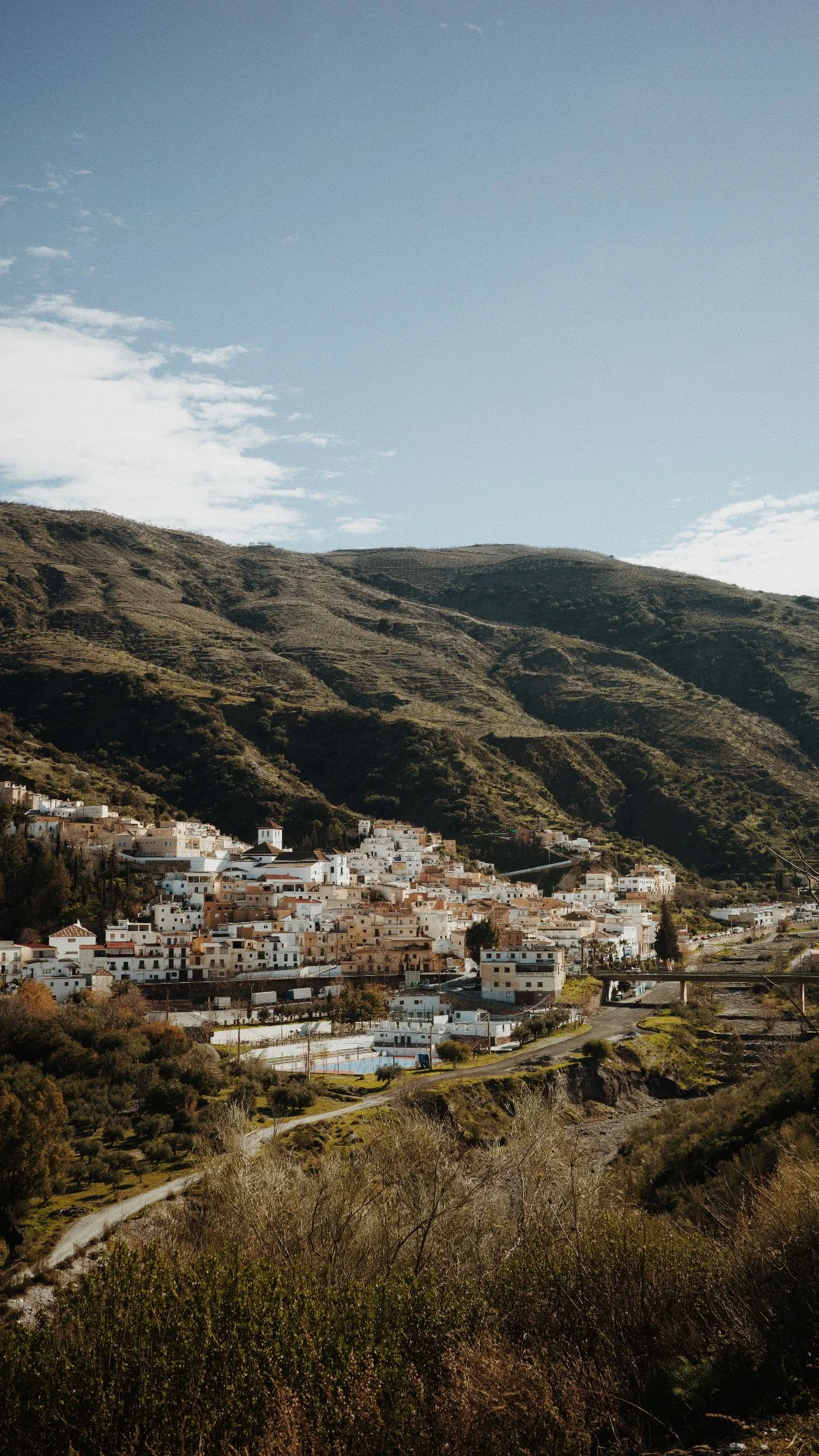 Vue d'un village blanc construit sur une colline, avec des montagnes en arrière-plan et un ciel bleu avec quelques nuages.