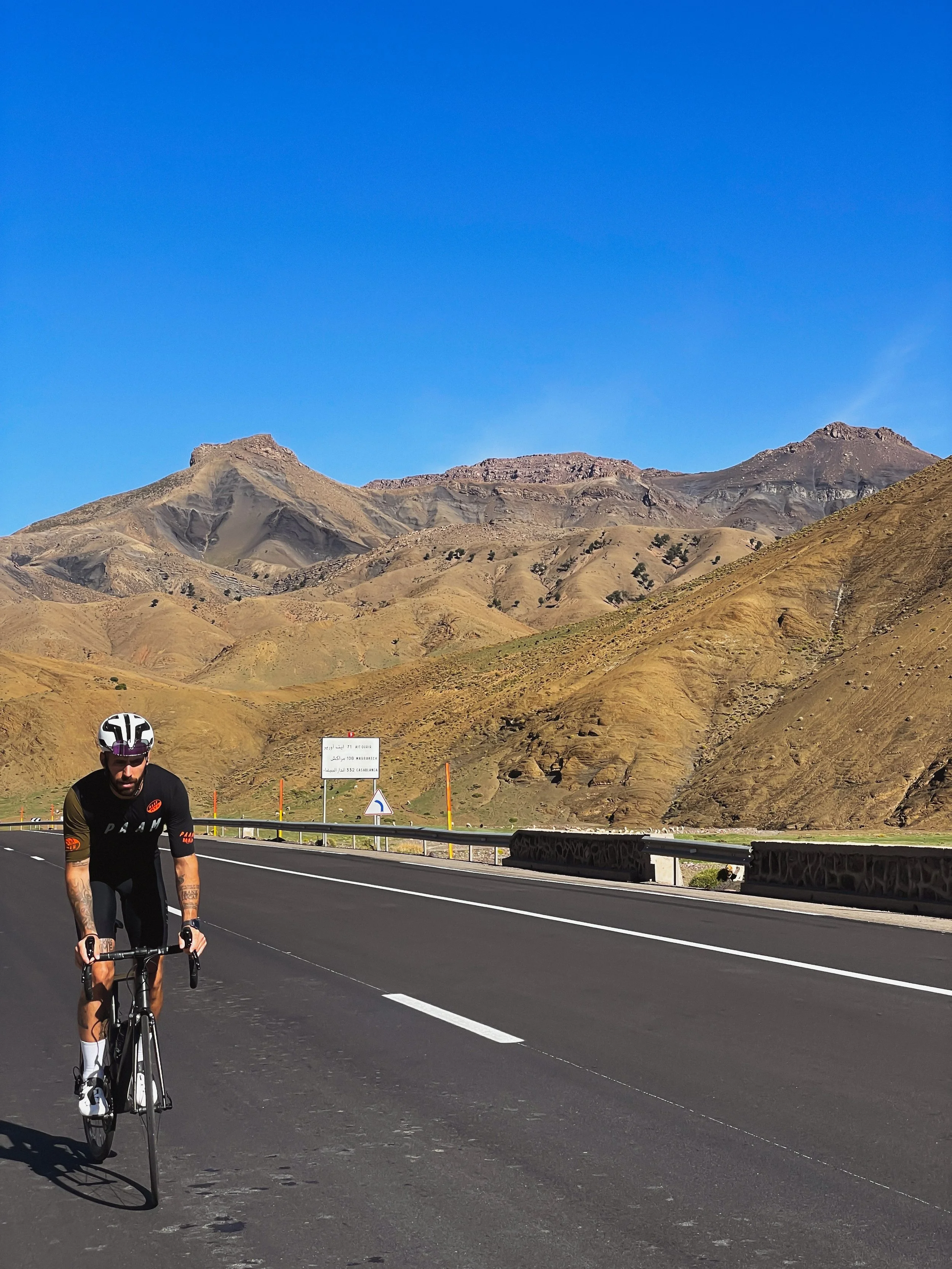 Un cycliste portant un casque noir sur une route pavée, avec un paysage de montagnes brunes sous un ciel bleu clair.