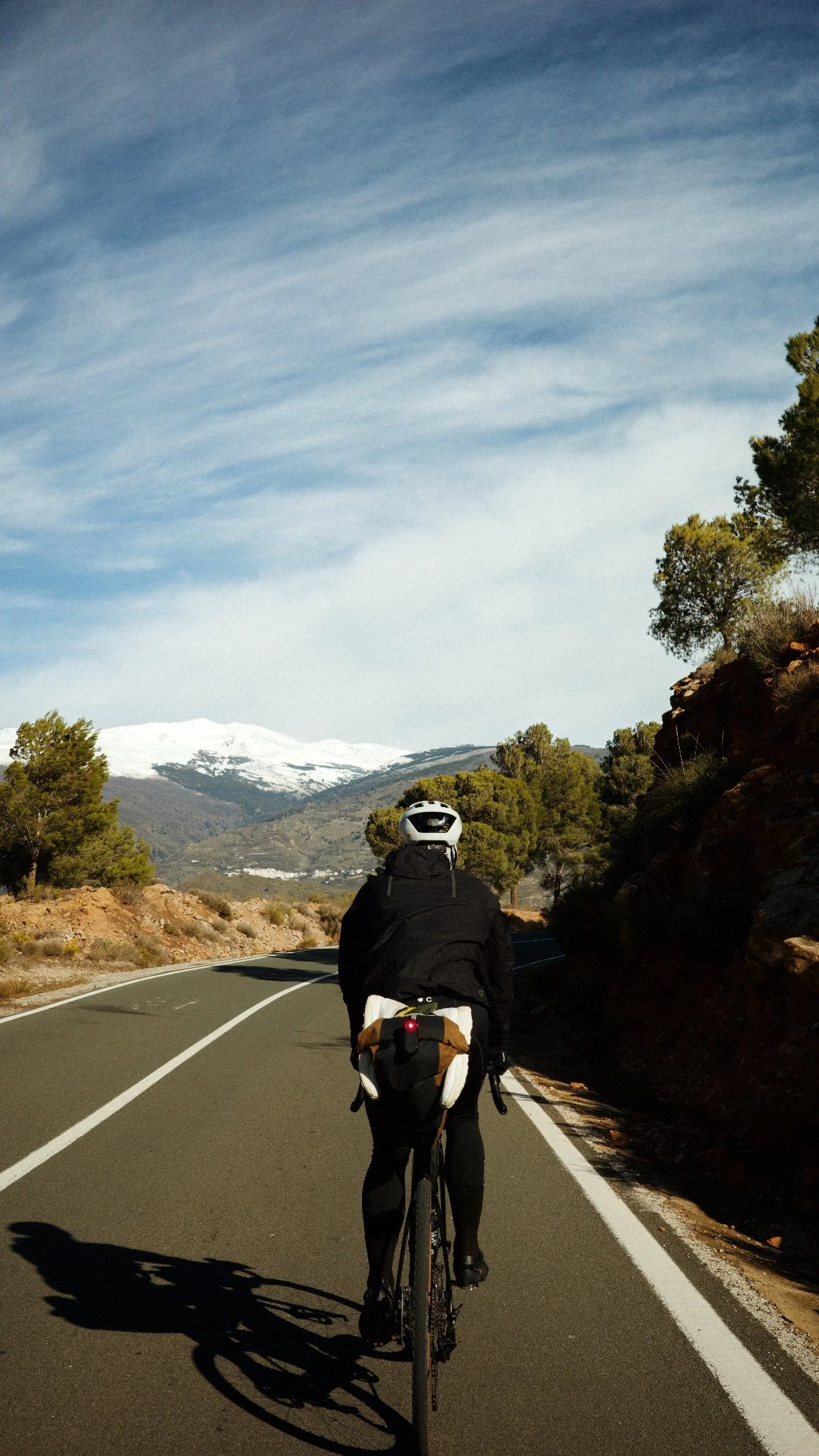 Une personne fait du vélo sur une route sinueuse dans un paysage de montagne avec des arbres et des montagnes enneigées à l'arrière-plan, sous un ciel bleu avec des nuages dispersés.