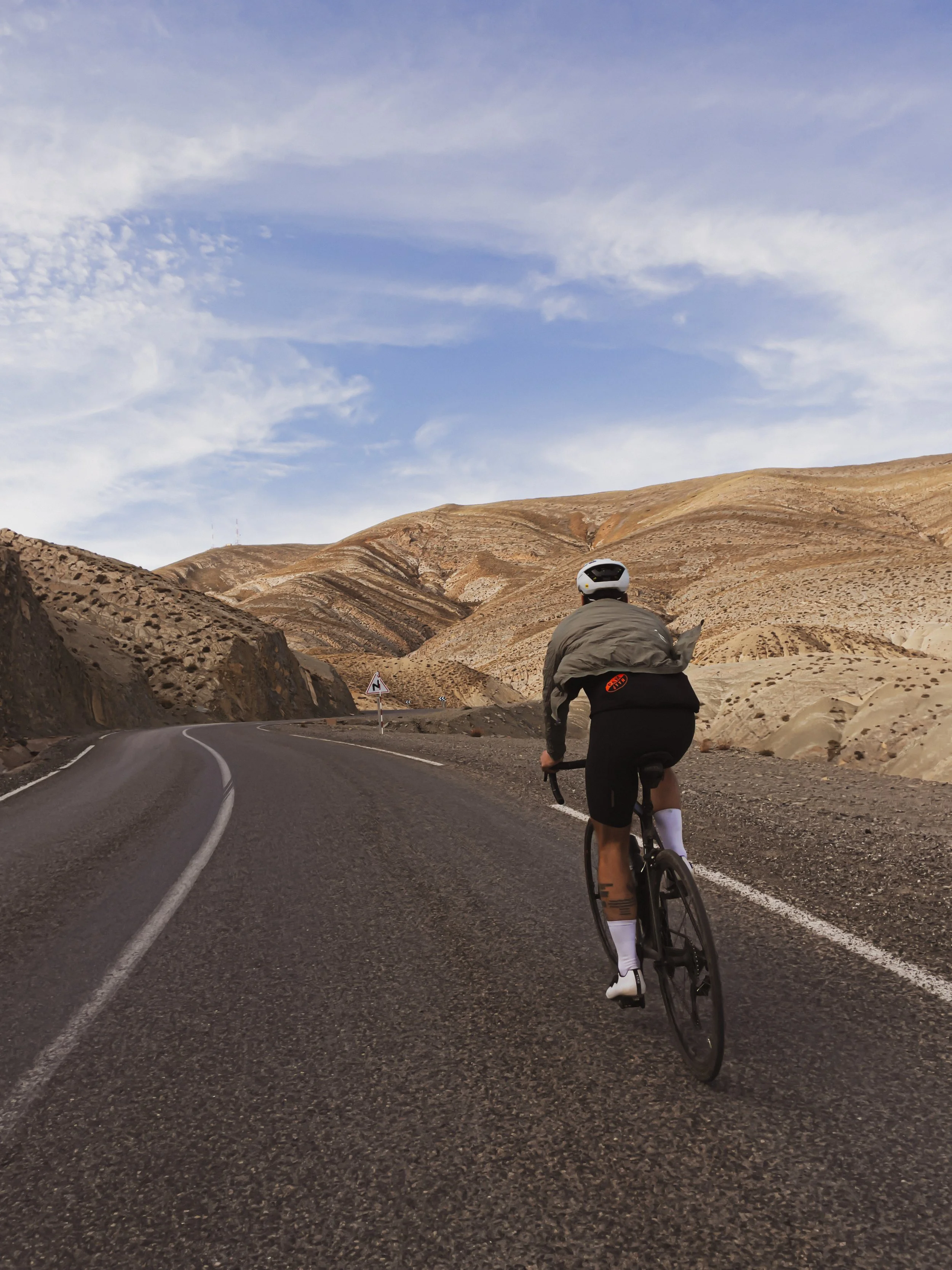 Un cycliste portant un casque traverse une route sinueuse dans un paysage désertique avec des collines rocheuses et un ciel clair.