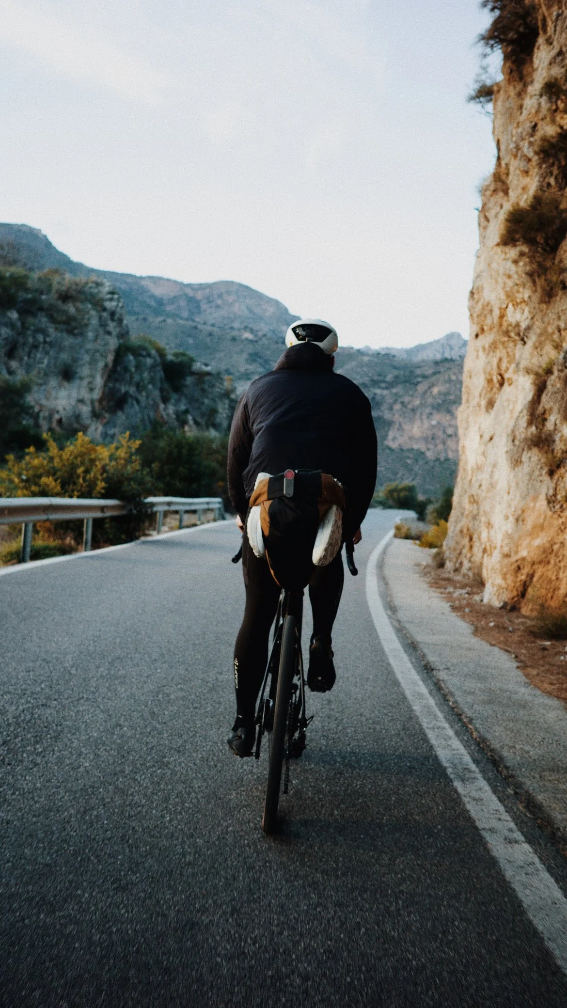 Un cycliste portant un casque, une veste noire et un sac à dos, pédale sur une route de montagne avec formations rocheuses et montagnes en arrière-plan.