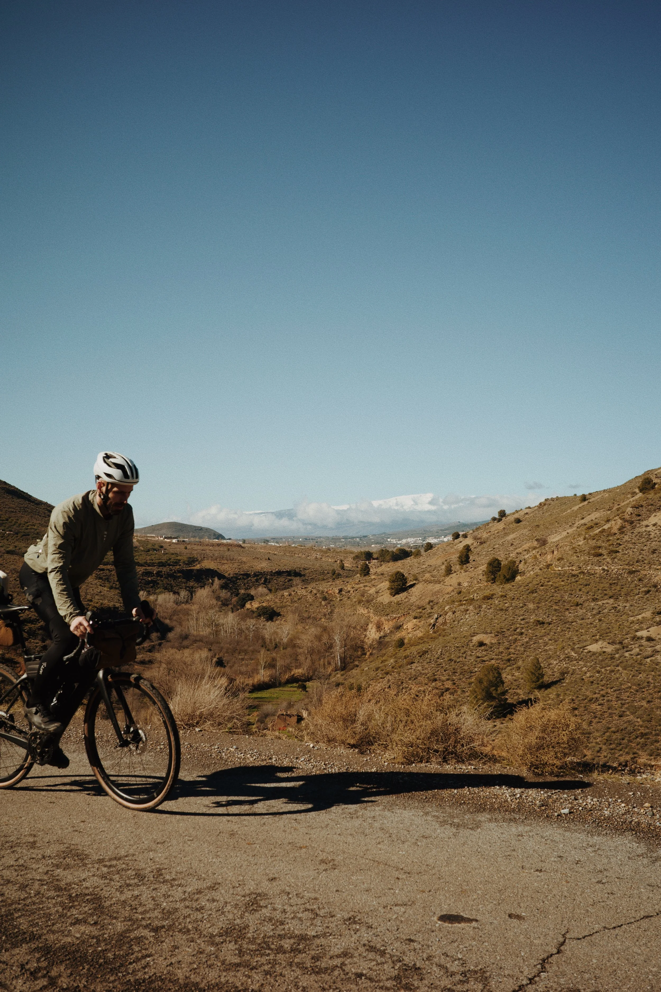 Un homme à vélo portant un casque, pédalant sur une route de campagne avec un paysage de collines arides, sous un ciel dégagé.