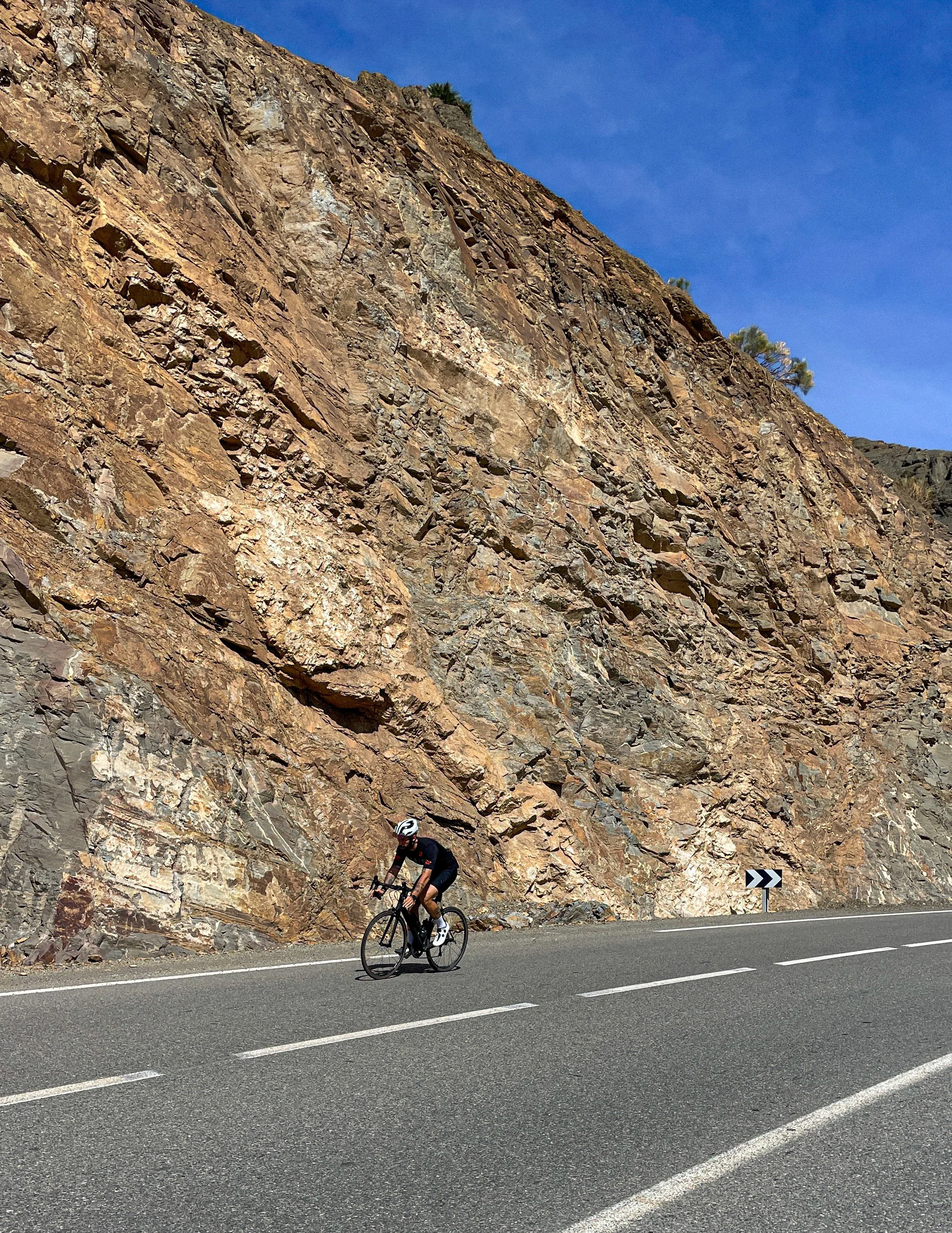 Un cycliste en pleine montée sur une route au pied d'une falaise rocheuse en terrain montagneux. Le ciel est clair et bleu.