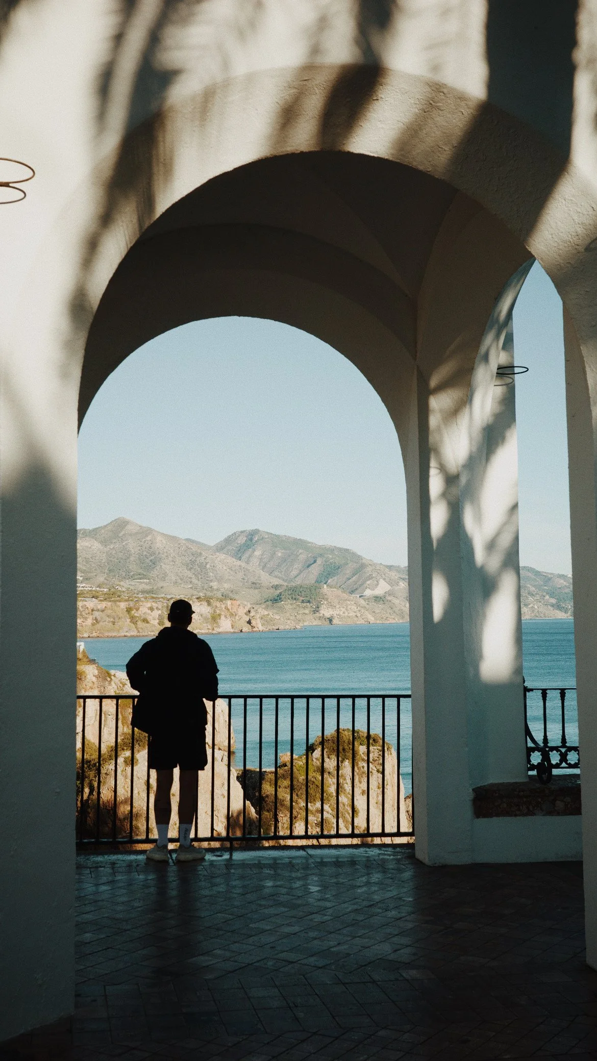 Une silhouette d'une personne regardant la mer à travers une arche blanche, avec des montagnes en arrière-plan et des ombres de branches d'arbres sur le mur.
