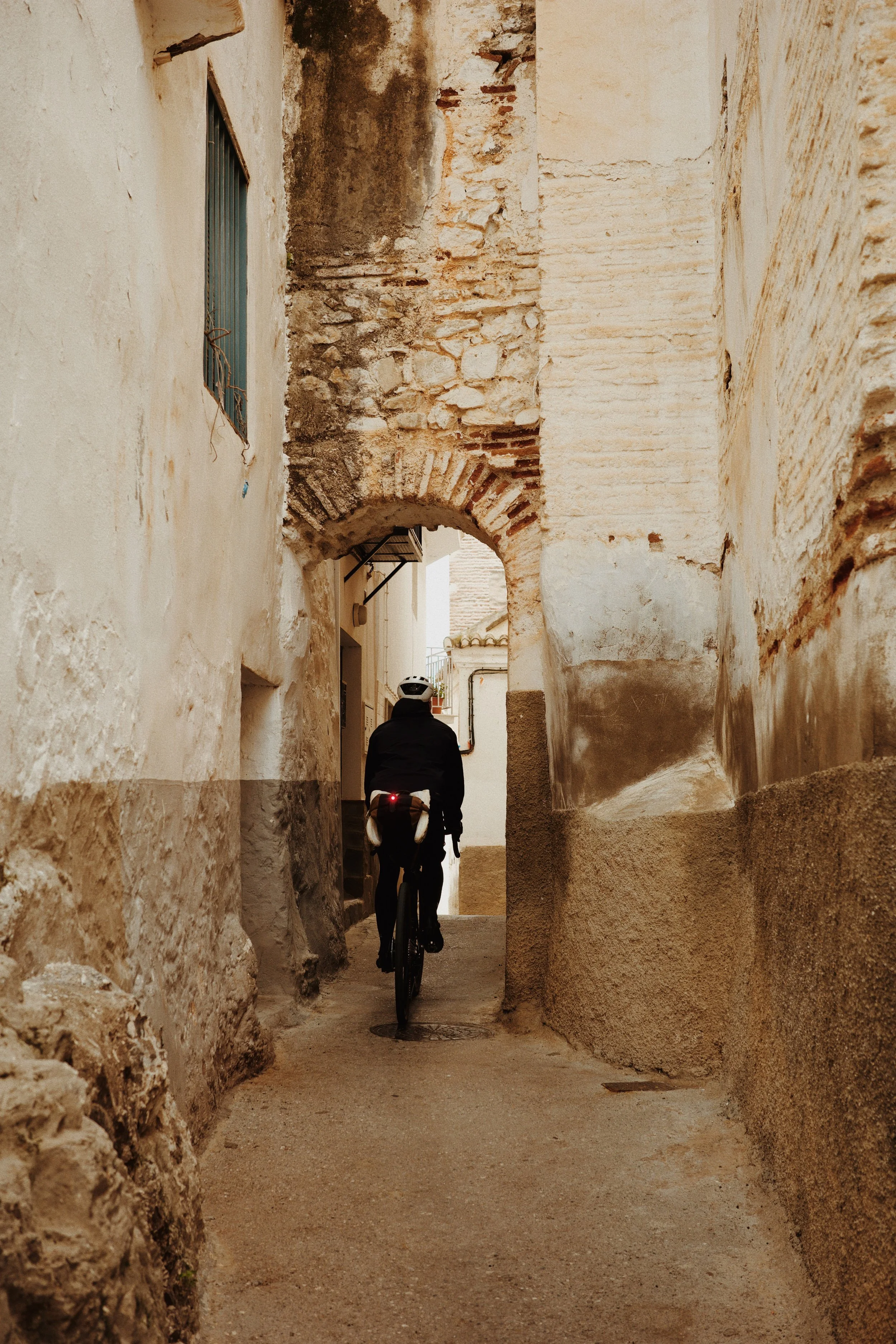 Un homme à vélo circule dans une ruelle étroite entourée de murs en pierre et en plâtre dans un village ancien, avec un arch en pierre à l'arrière.